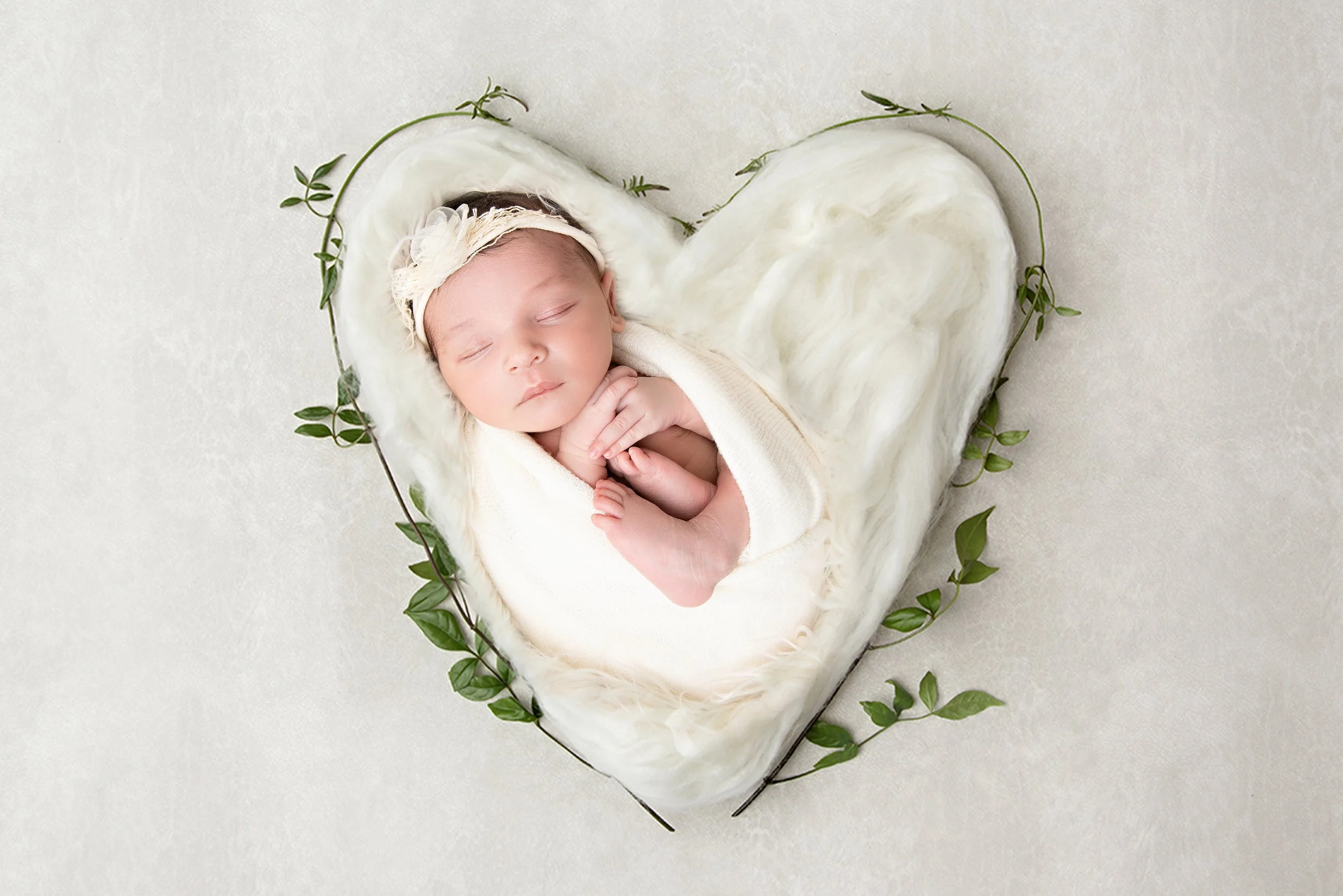 Sleeping baby girl wrapped in a white blanket, wearing a headband, lying on a fur-like surface surrounded by greenery, on a white background.