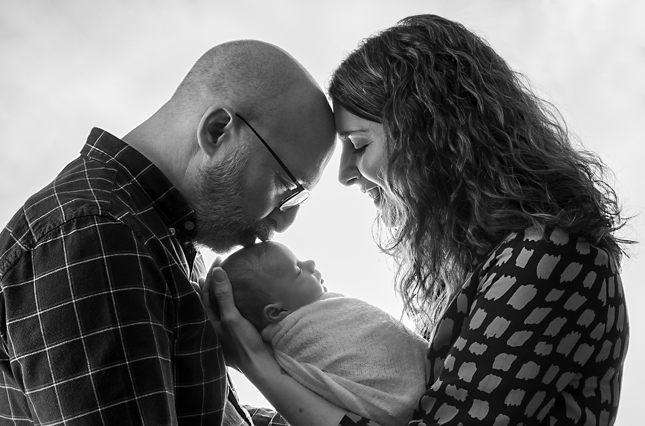 A black and white photo of a family moment where a man and woman are touching foreheads, holding a newborn baby between them. The man wears glasses and a checkered shirt, the woman has curly hair, and the baby is wrapped in a blanket.