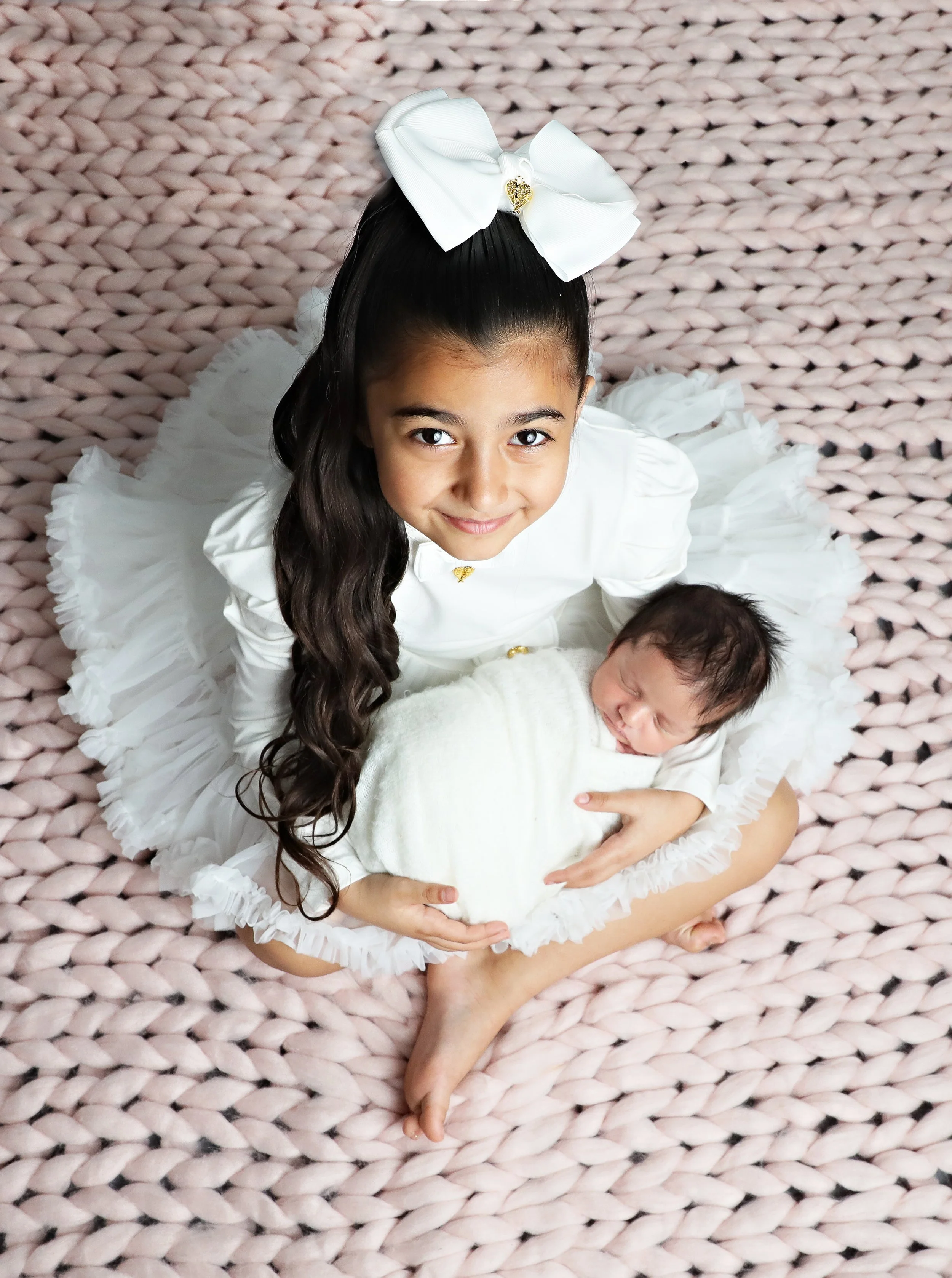 A young girl with long dark hair smiling, wearing a white dress with ruffled sleeves and a large white bow in her hair, holding a small white puppy, on a pink knitted blanket.