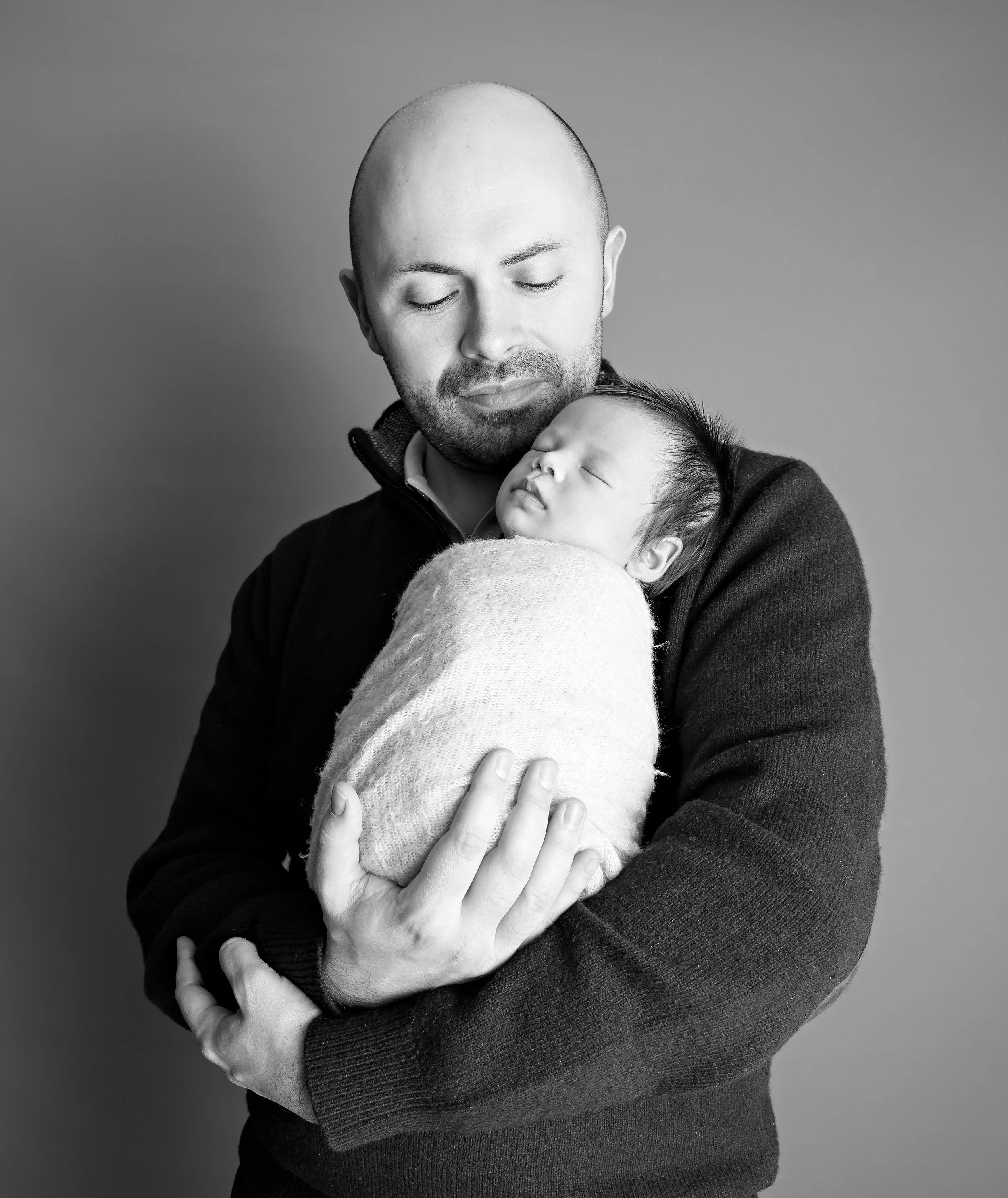 A black-and-white photograph of a man with a shaved head and beard holding a sleeping baby wrapped in a blanket, with both closing their eyes.