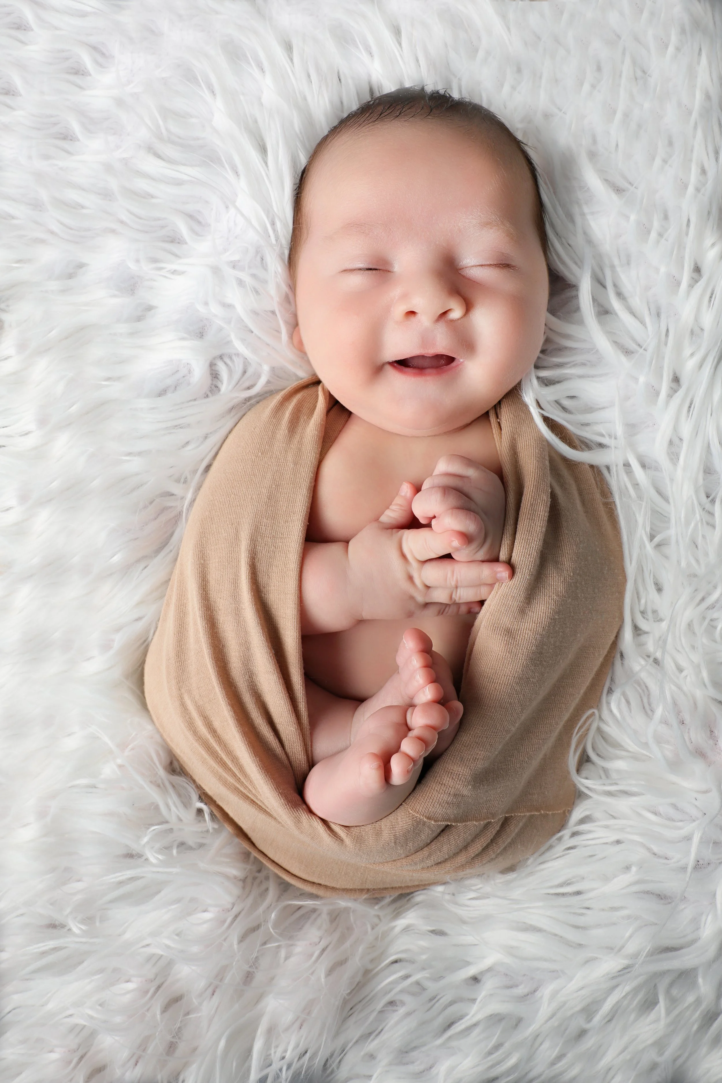 A smiling baby lying on a white fluffy blanket, clasping hands together, wearing a tan wrap.