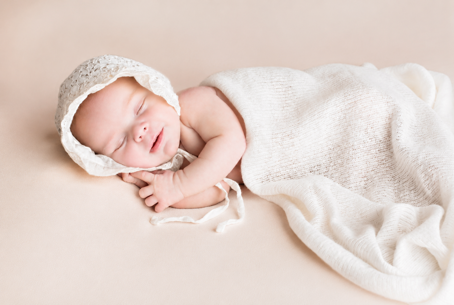 A sleeping baby lying on a beige surface, wearing a white lacy bonnet and swaddled in a white, loosely knitted blanket.