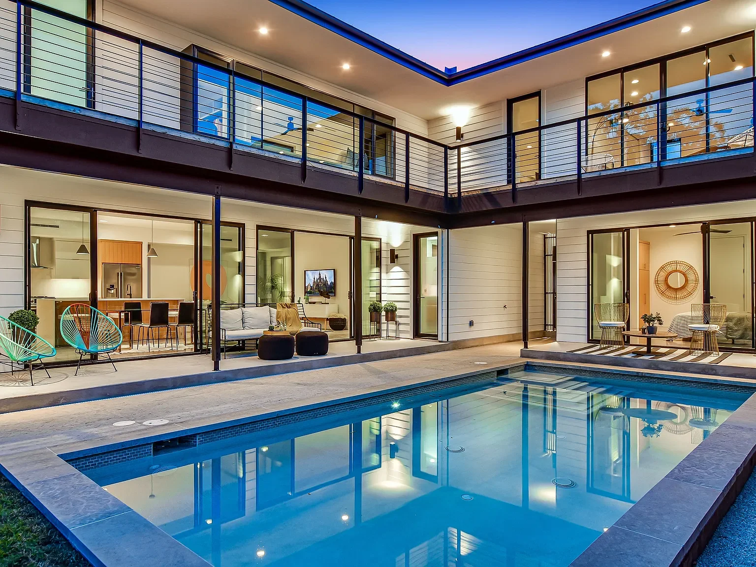 Modern residential backyard with swimming pool, outdoor seating area, and large glass sliding doors leading into the house, illuminated indoors during evening.