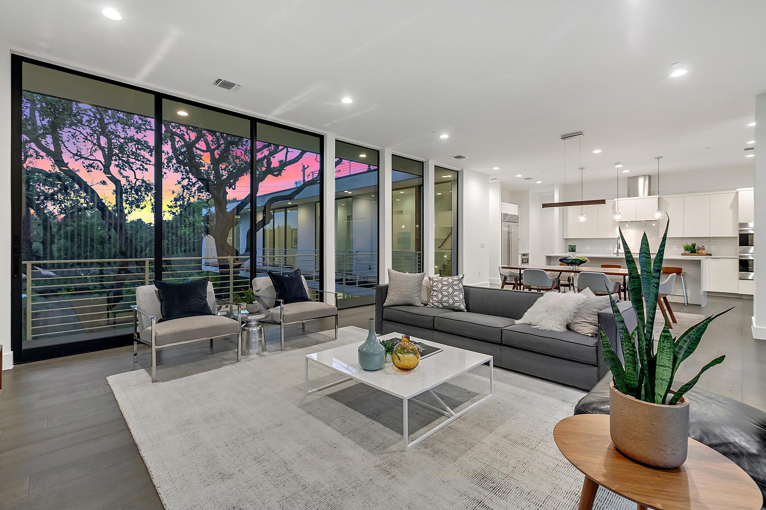 Modern living room with large windows showing a colorful sunset sky, furnished with a black sofa, white coffee table with decorative vases, beige armchairs, and various throw pillows, along with indoor plants and minimalist decor.