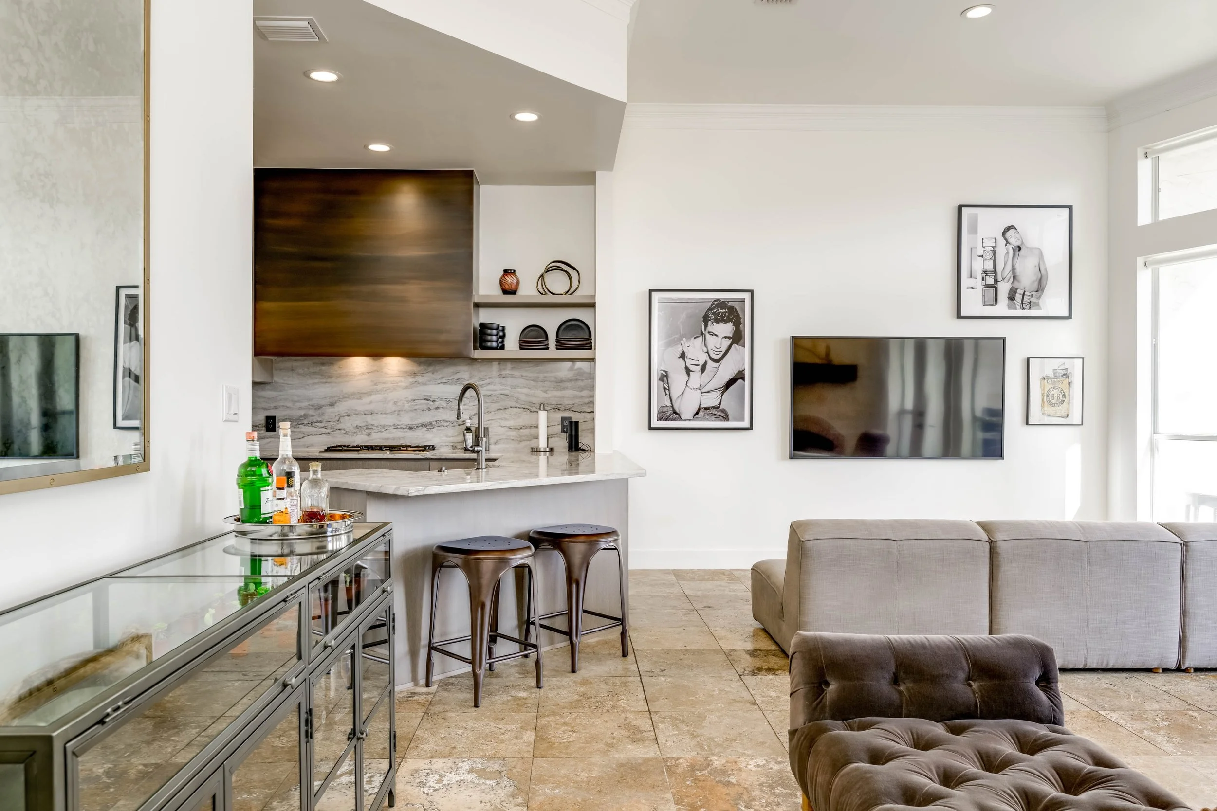 A living room and kitchen area with a marble kitchen island, framed photographs on the white walls, a large flat-screen TV, and a black tufted armchair. There is a tray with bottles and glasses on a glass display case, and bar stools with wooden seats and metal legs at the kitchen counter. Large windows with white blinds let in natural light.