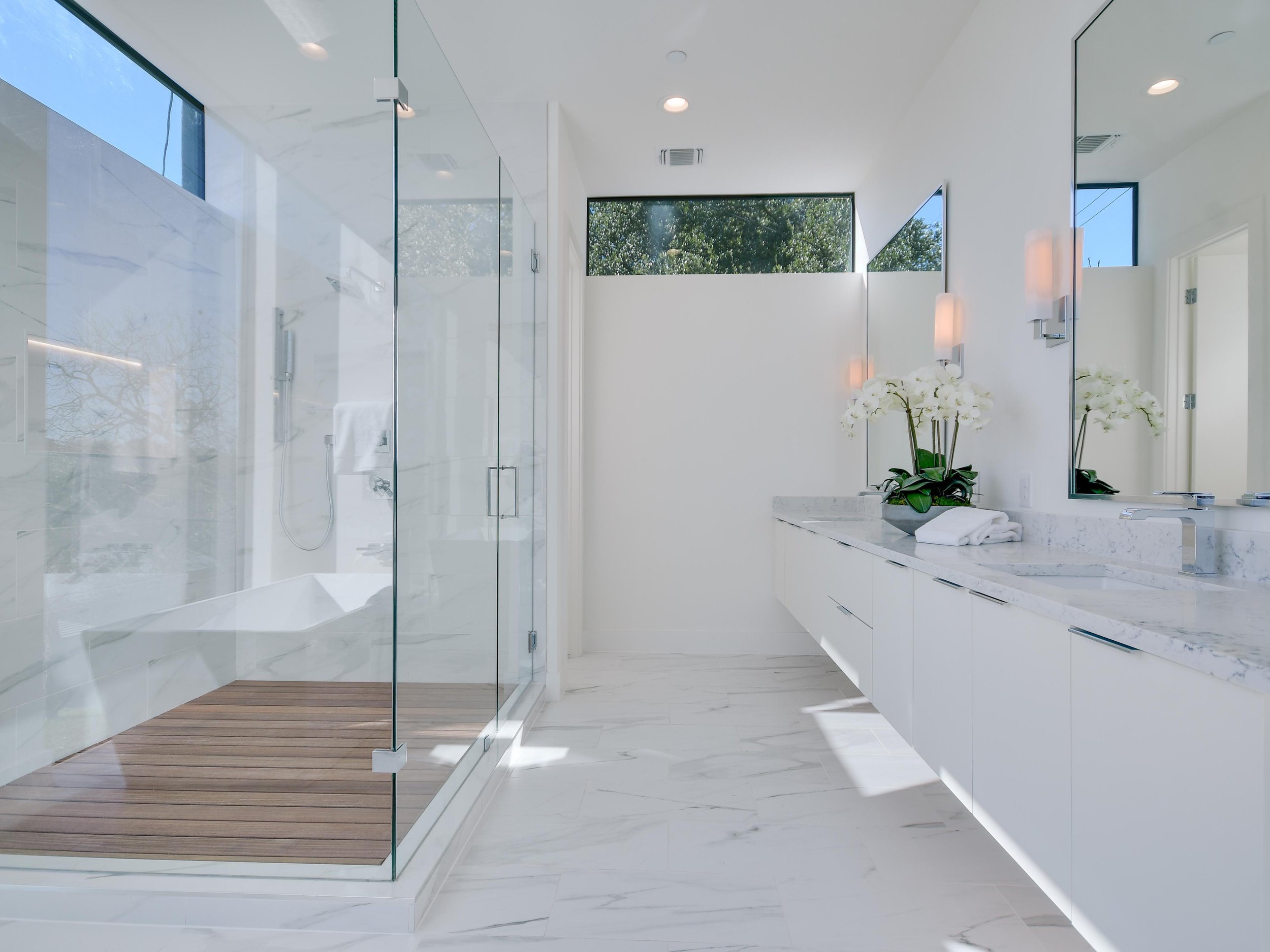 Modern bathroom with white marble walls and floors, a glass shower stall, a white vanity with a large mirror, and large windows showing trees outside.