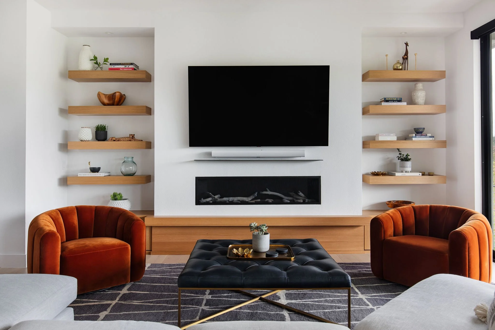 Living room with white walls, orange velvet armchairs, a black tufted ottoman with gold legs, and a black and white geometric rug. A white fireplace is centered on the wall, with a flat-screen TV mounted above and a soundbar beneath. Built-in wooden shelves on either side hold decorative items such as books, vases, plants, and ornaments.