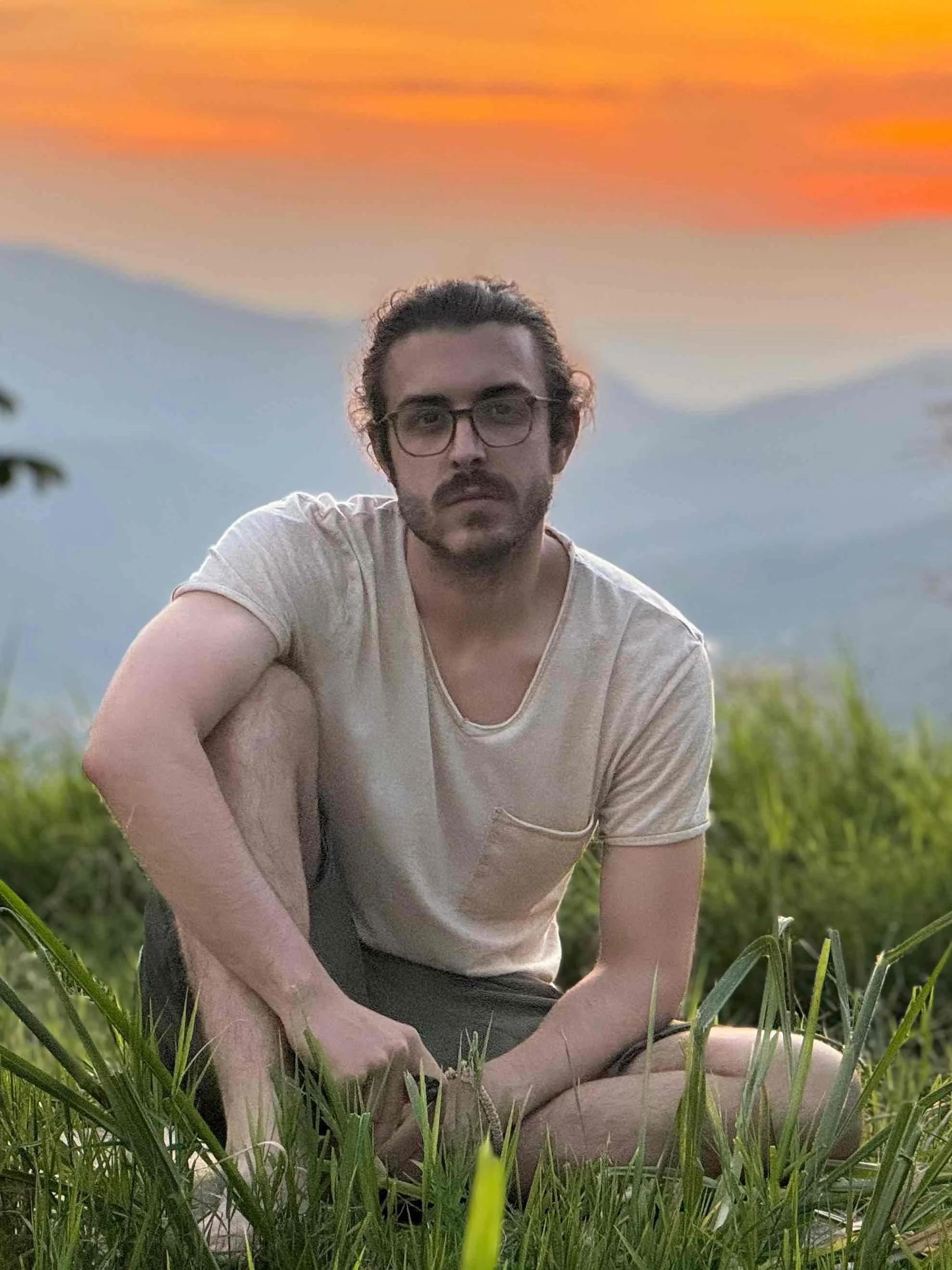 A man with glasses, curly hair, and a beard sitting in a grassy field during sunset, looking at the camera.