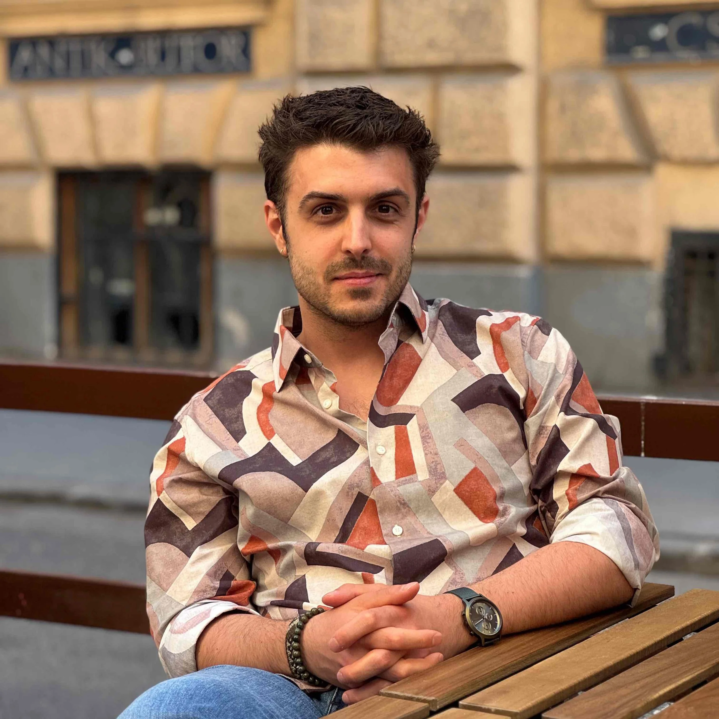 A young man with dark wavy hair, wearing a multicolored patterned shirt, sitting at a wooden table with his arms crossed, and looking directly at the camera. Background includes a brick wall and a bookshelf.