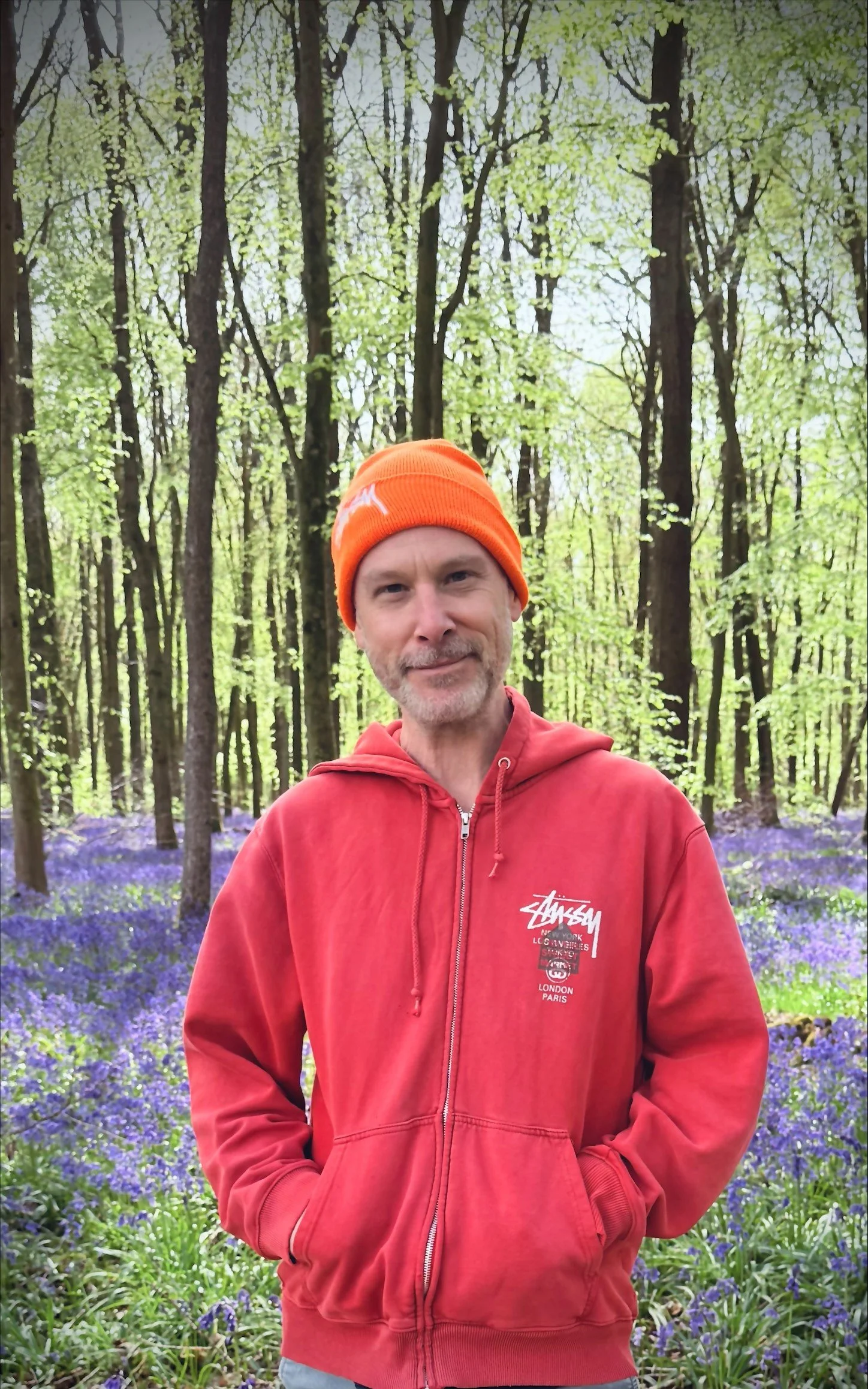 A man with a slight beard and mustache, wearing an orange beanie and red hoodie, standing in a forest with tall trees and purple flowers on the ground.
