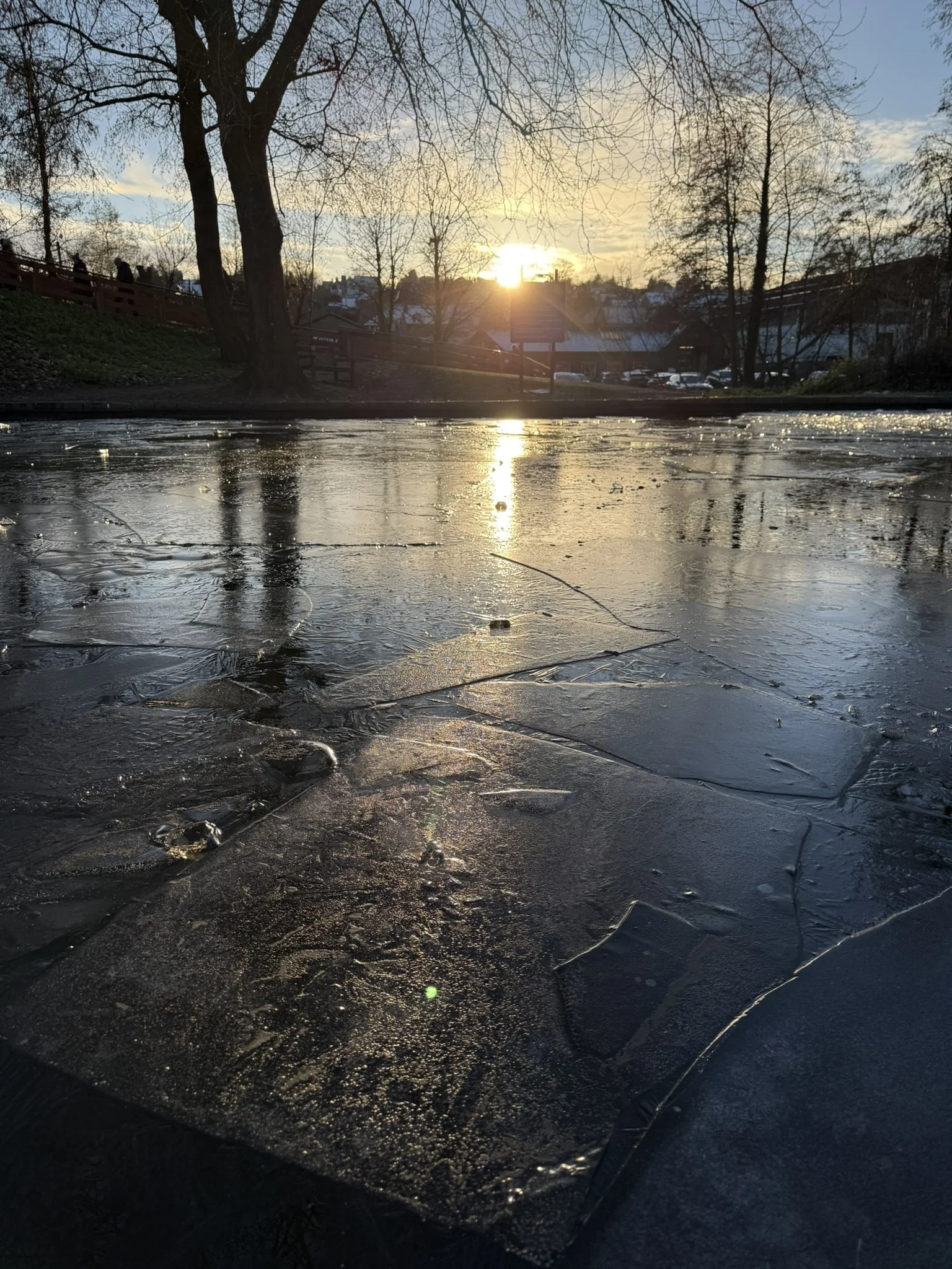 The image captures a scenic view of a sunset over a frozen body of water, with ice patches and cracks visible on the surface. In the background, trees and buildings are silhouetted against the colorful sky, with the sun partially hidden behind the trees.