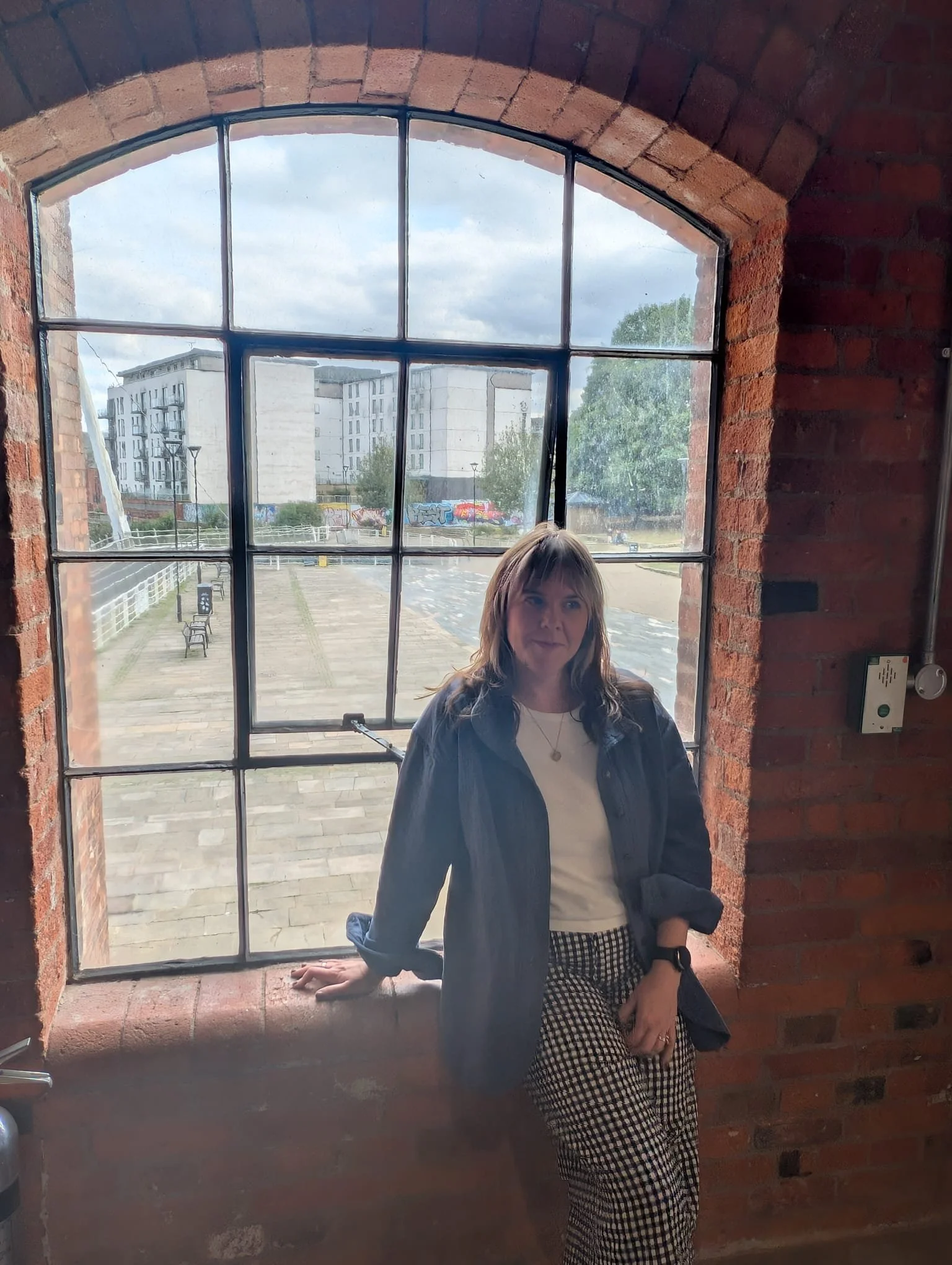 A woman with brown hair and a black jacket standing inside a building, looking out a large brick-framed window with metal grids, with an urban outdoor scene of white apartment buildings and some graffiti visible in the background.