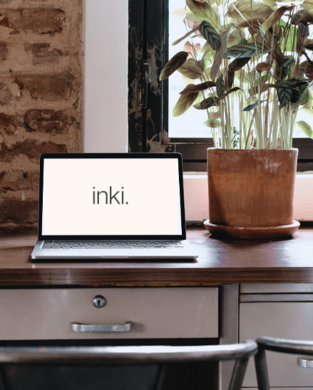 A laptop on a wooden desk displaying the word 'inki.', next to a large potted plant, with a window and brick wall in the background.