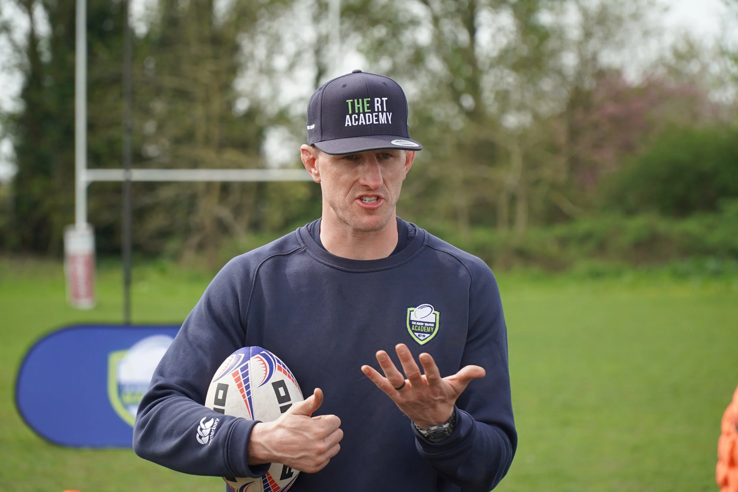 A man wearing a black cap with the logo 'The RT Academy' and a dark sports jacket, holding a rugby ball, appears to be coaching or instructing during an outdoor rugby training session.