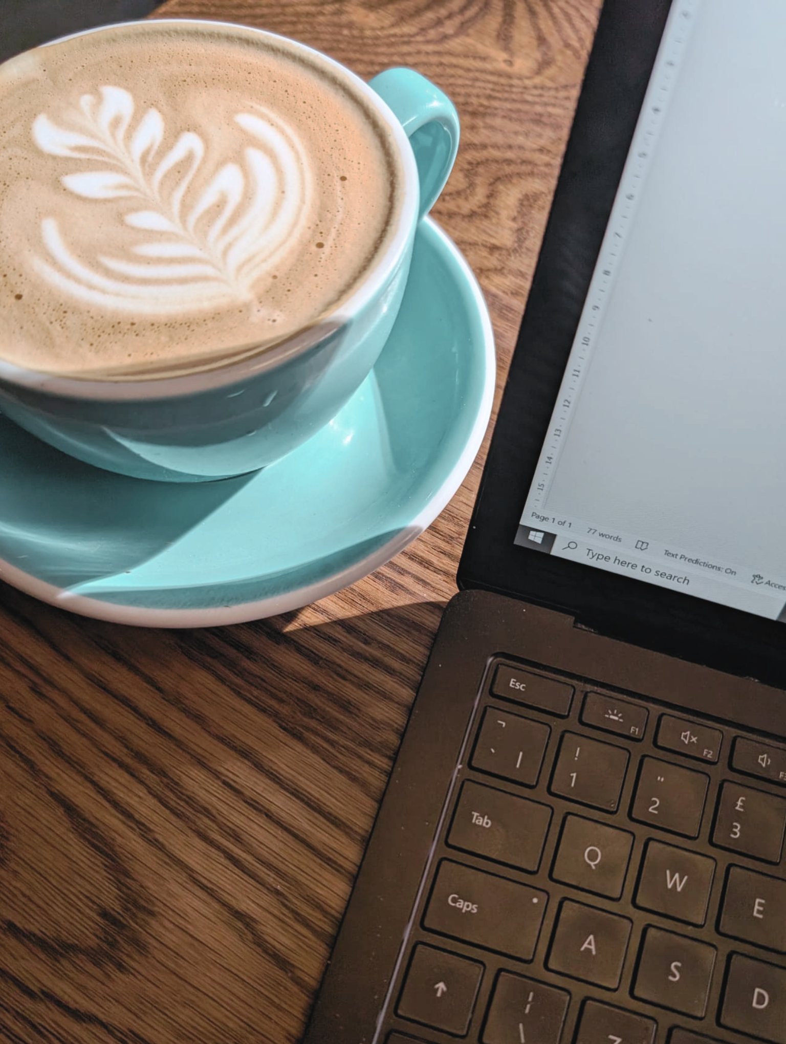 A cup of coffee with latte art on top, sitting on a tray next to a laptop on a wooden table.