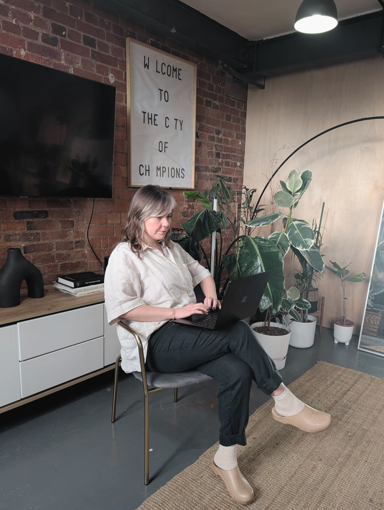A woman sitting on a chair in a modern interior space, working on a laptop. The space features a brick wall with a welcoming sign, potted plants, and a minimalist sideboard with books and decorative items.