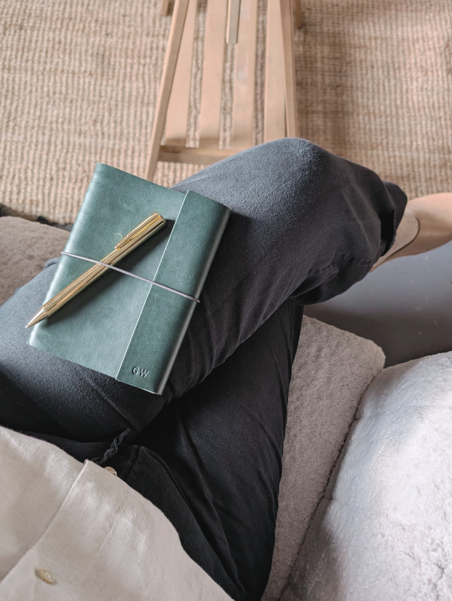 A person sitting with legs crossed, wearing black pants and a beige coat, holding a green notebook and gold pen. The person is seated on a carpeted floor near a wooden bench.