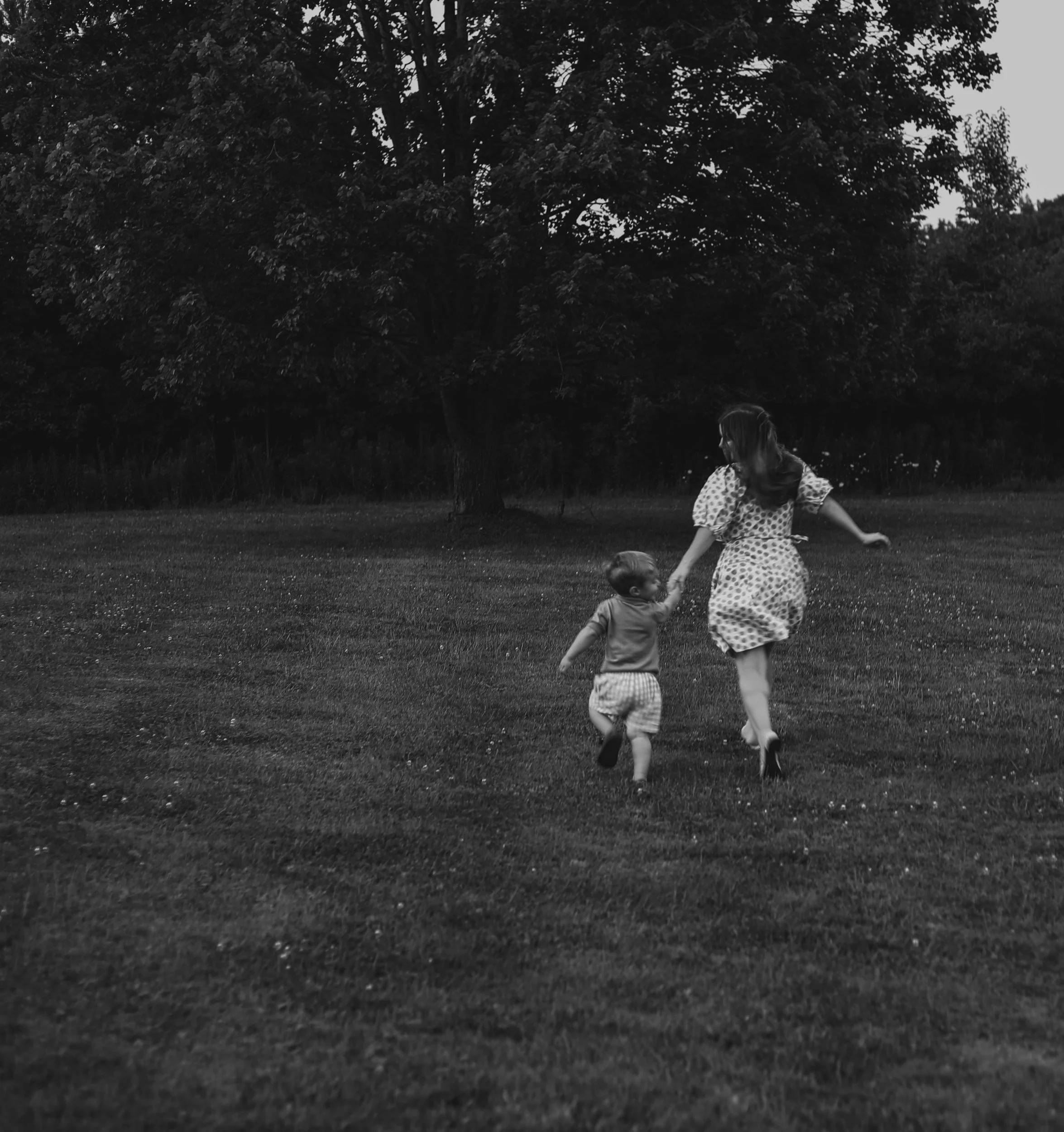 A woman and a young child are playing in a grassy park area near a large tree, with other trees visible in the background; the scene is in black and white.