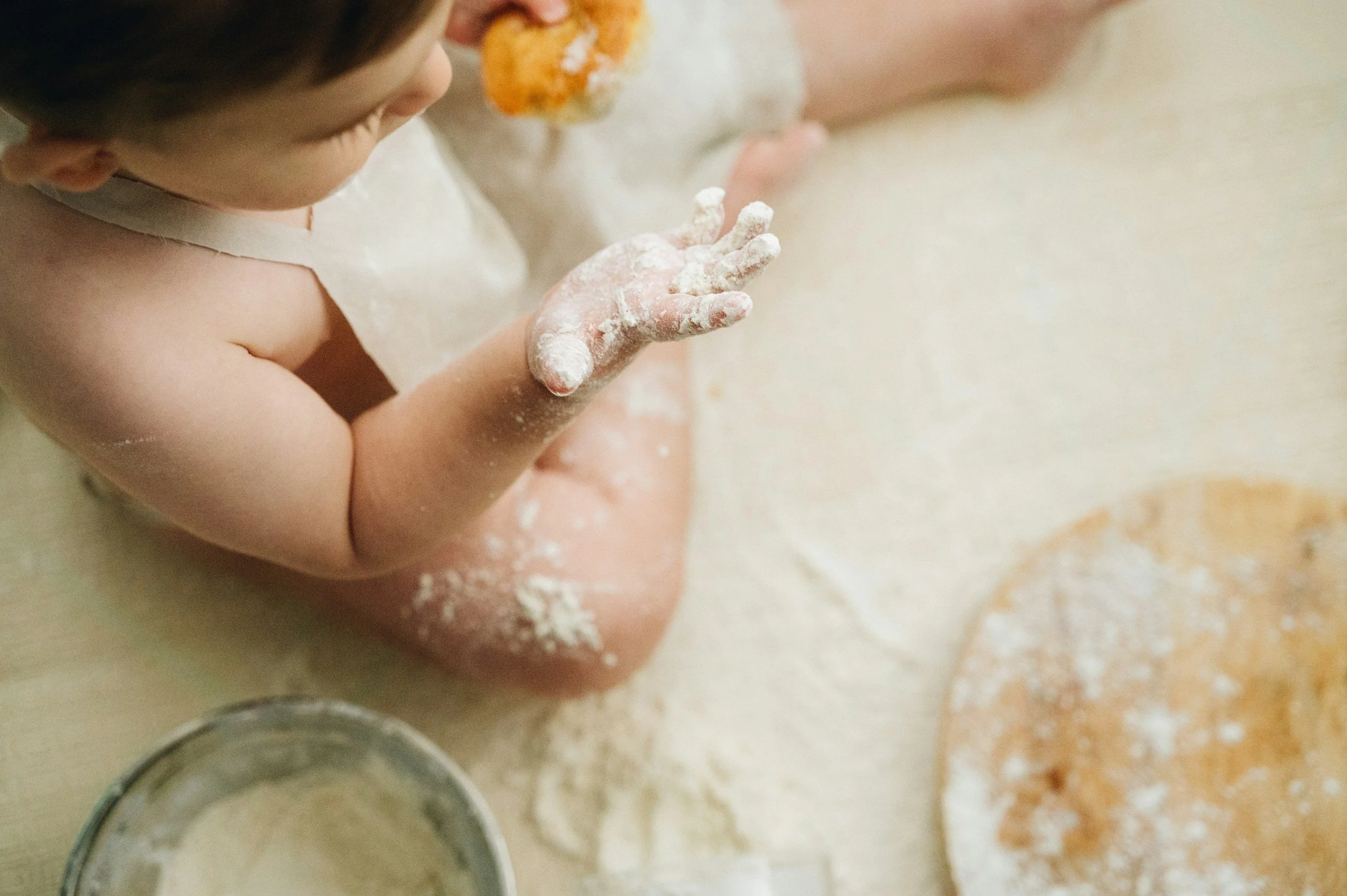A young child with short brown hair is sitting on a countertop, covered in flour, with a piece of breaded food near their face. There are bowls and spilled flour on the countertop.