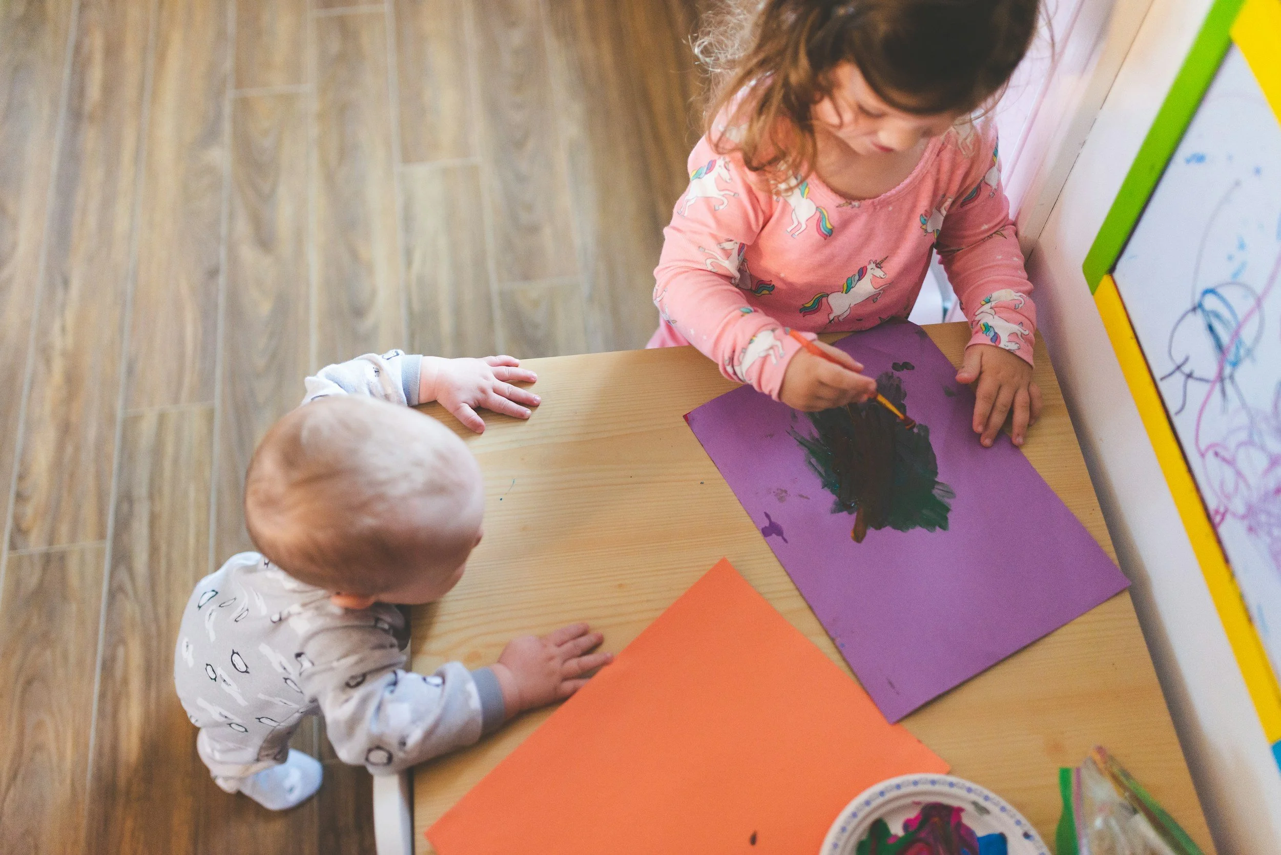 A young girl with curly hair wearing a pink shirt with unicorns, painting on purple paper at a wooden table, while another child in pajamas watches.