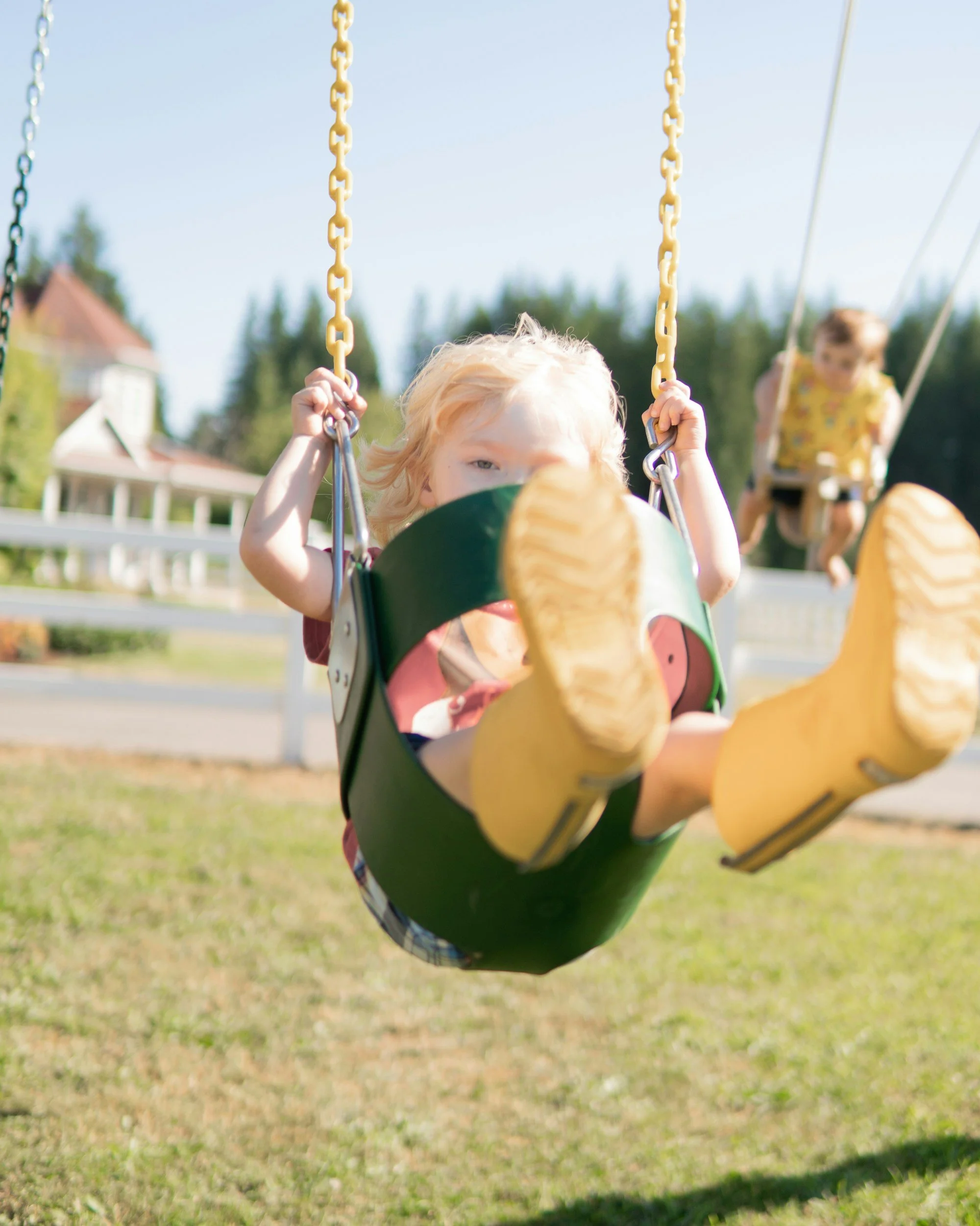Child on a swing in a park with a house and trees in the background, facing the camera and holding the chains of the swing.