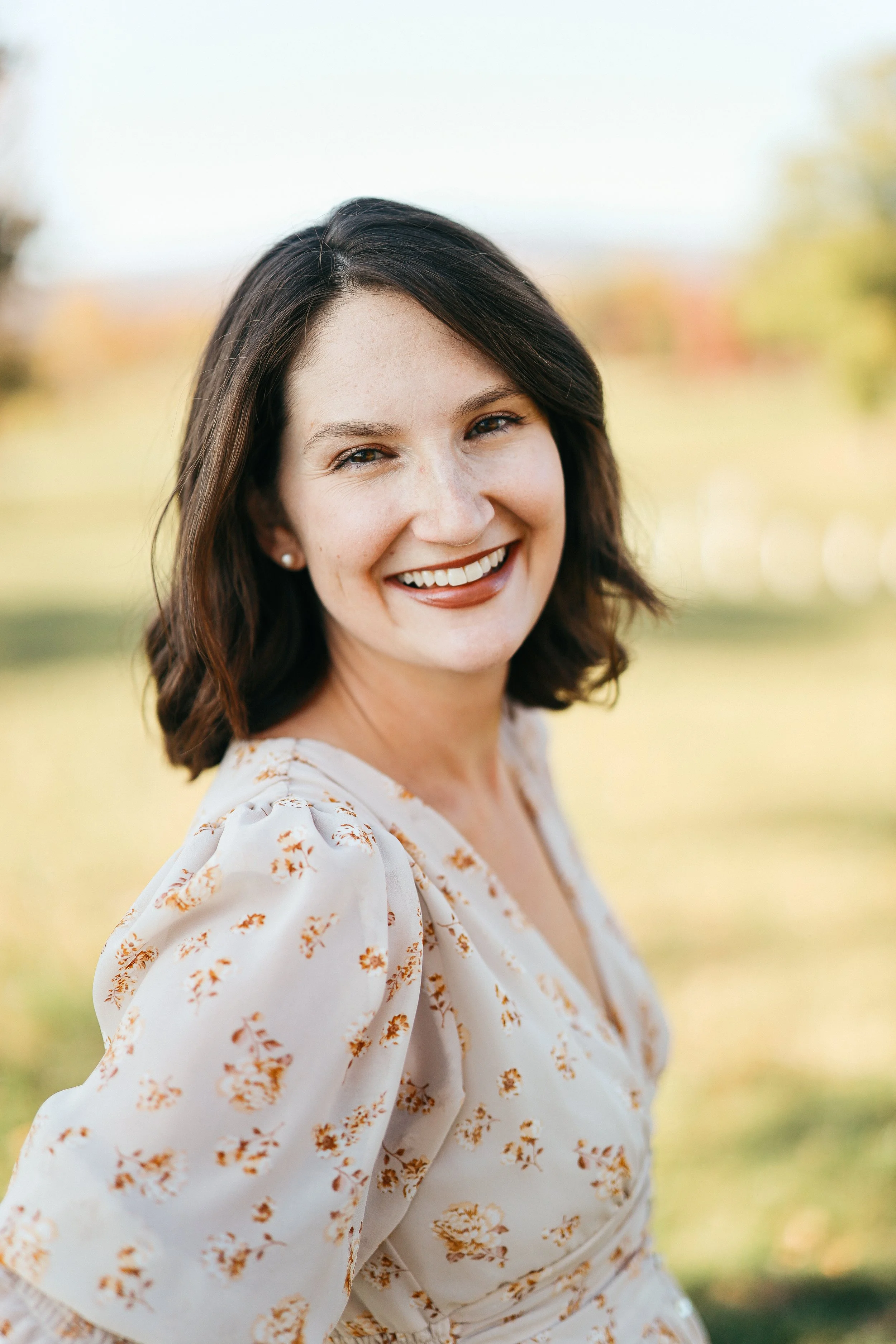 A woman with shoulder-length dark hair smiling outdoors with blurred trees and sky in the background, wearing a white blouse with orange floral print.