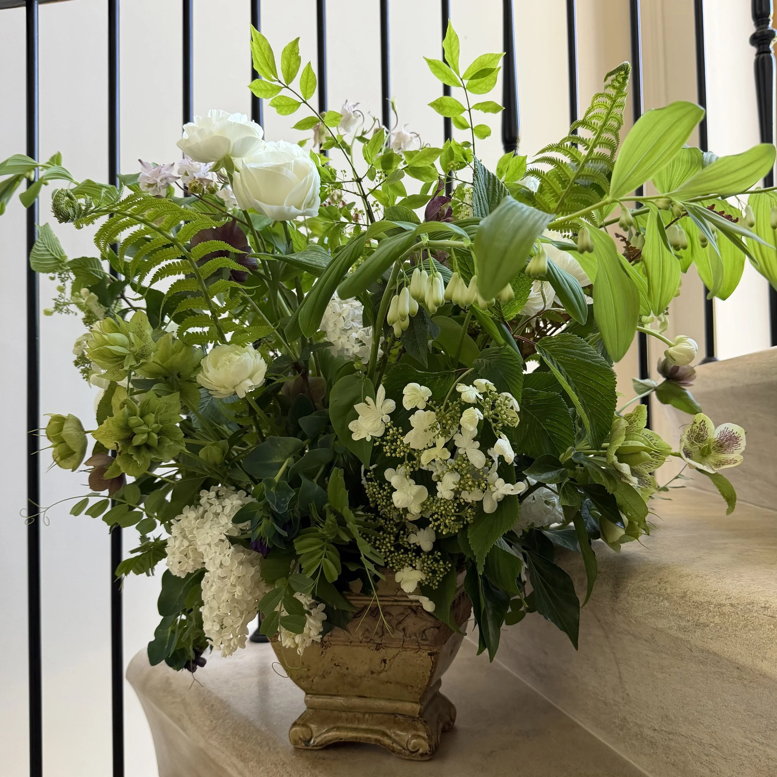 A floral arrangement in a stone-like decorative vase, containing white roses, green leaves, and various other white and purple flowers, placed on a beige countertop with a black metal railing in the background.