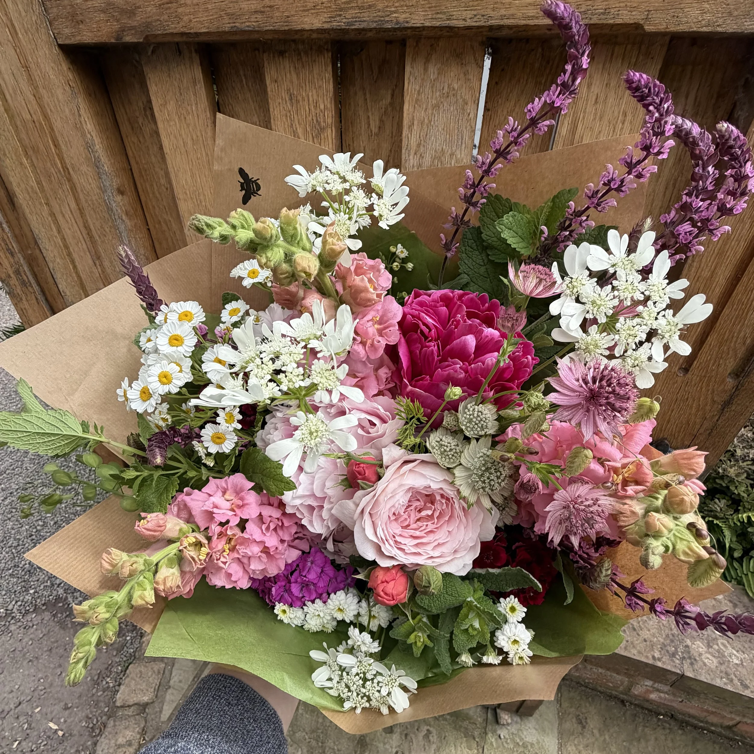 A colorful bouquet of mixed flowers including white daisies, pink peonies, purple lavender, and other blooms wrapped in brown paper, placed on a wooden surface outdoor.