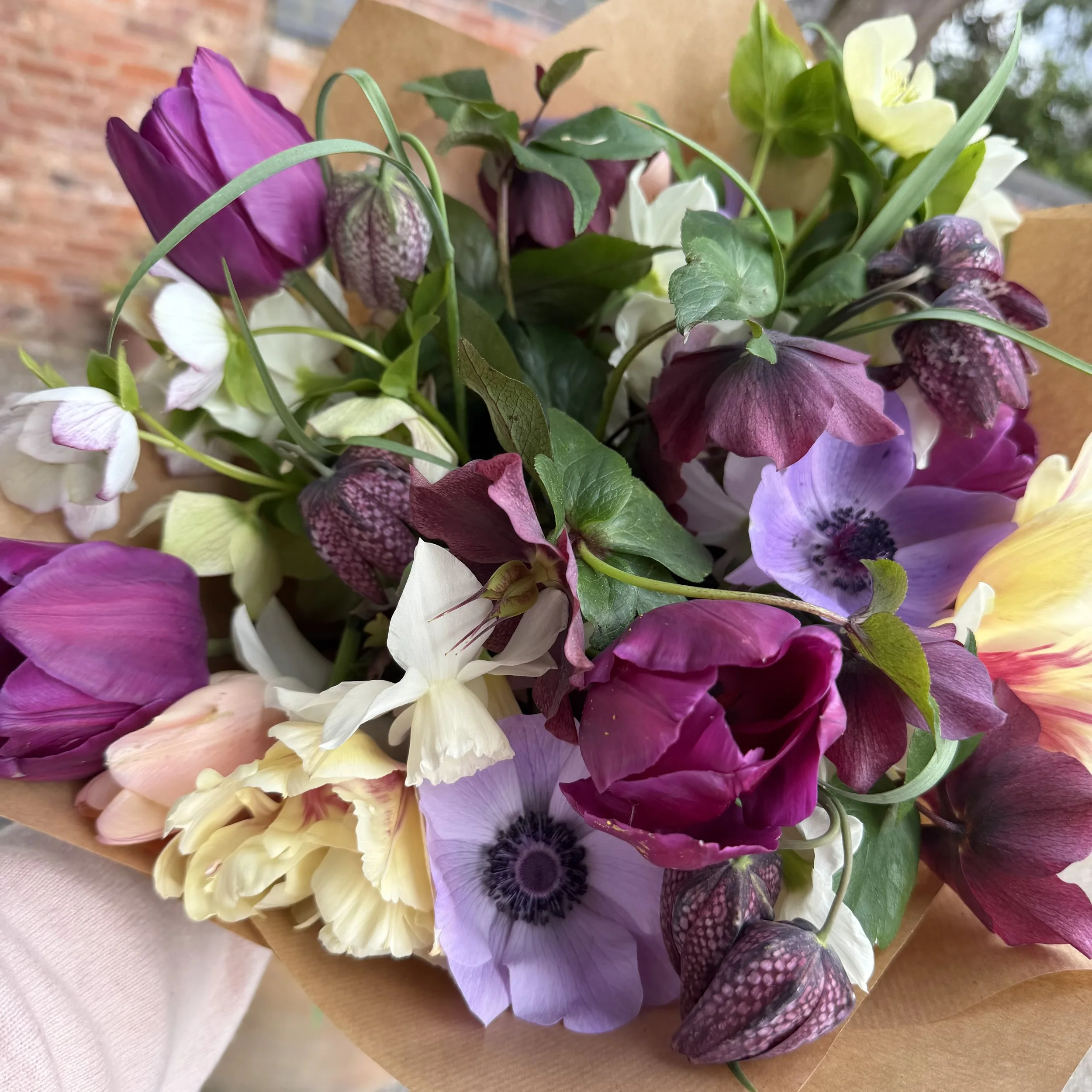 A colorful bouquet of assorted flowers including purple, white, yellow, and burgundy blooms wrapped in brown paper.