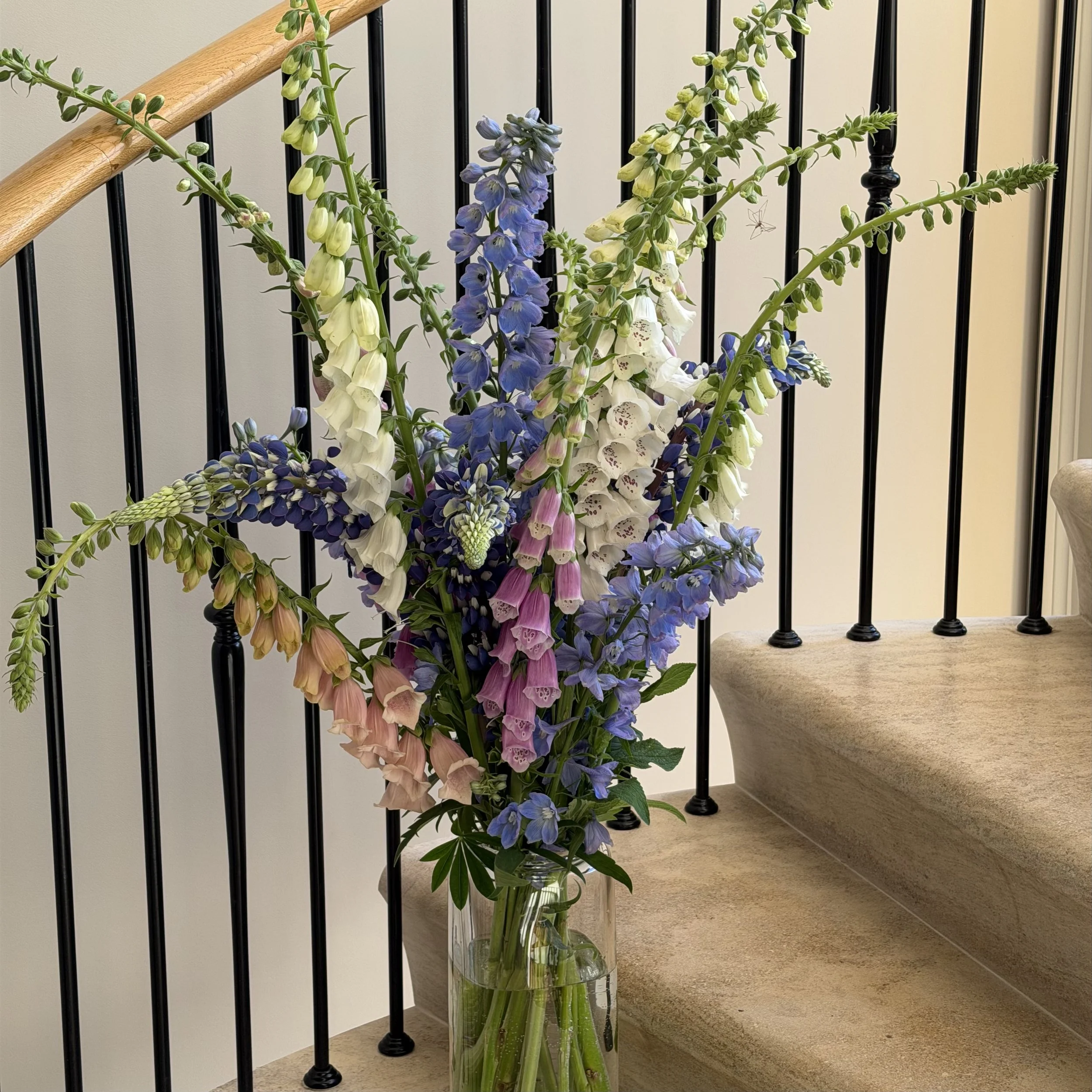 A tall glass vase filled with mixed tall flowers, including purple delphiniums, white and pink foxgloves, and blue and purple other flowers, placed beside a staircase with black metal railings and a wooden handrail, on a beige carpeted floor.