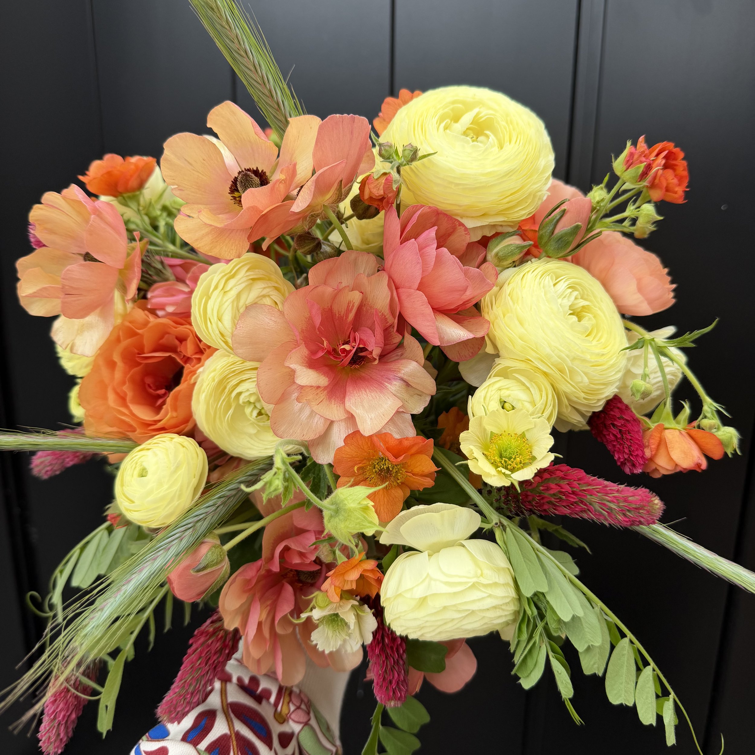 A colorful floral bouquet with yellow, pink, orange, and red flowers, accented with green leaves and grasses, set against a dark background.