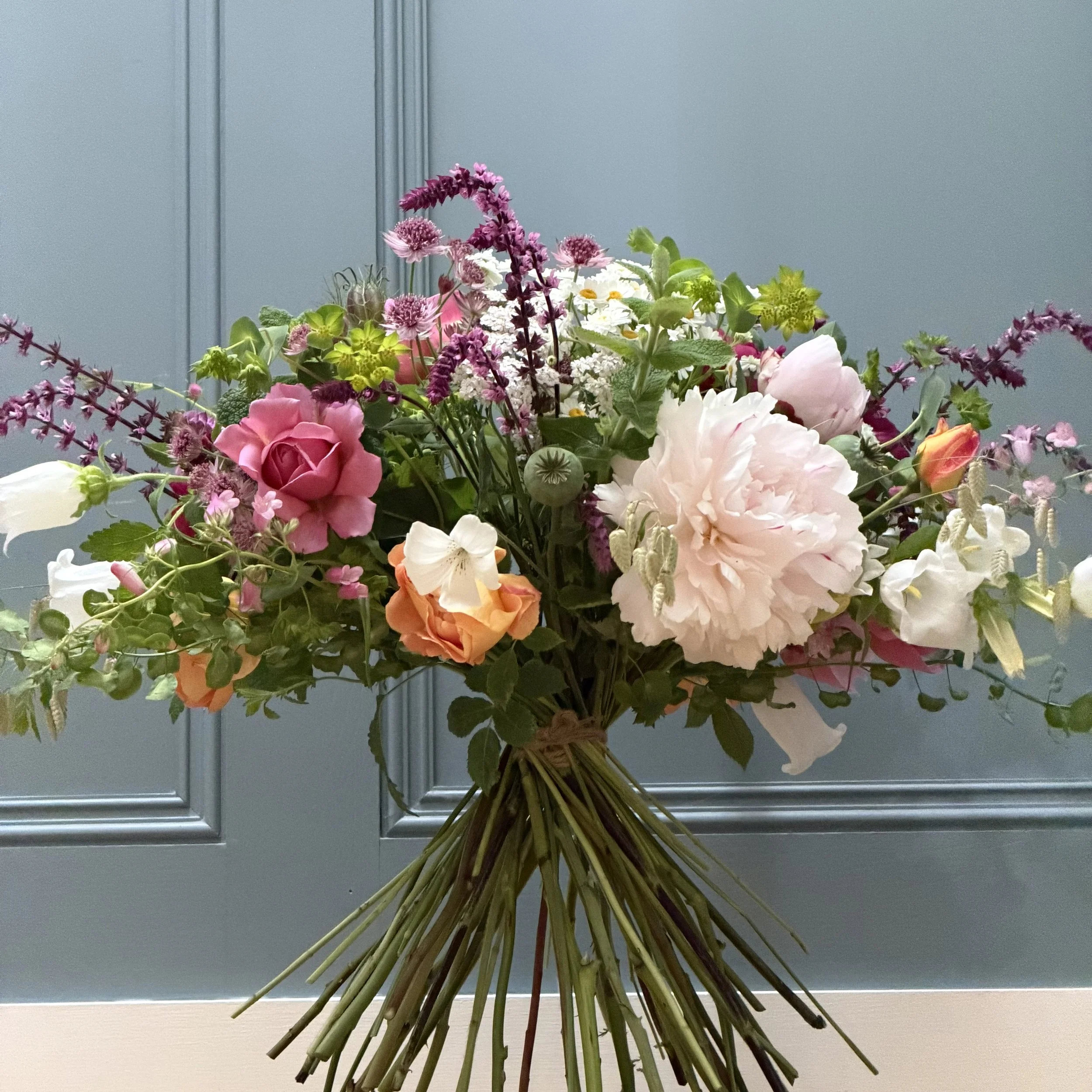 A bouquet of various colorful flowers including roses, daisies, and other blooms, arranged with green foliage, placed against a light blue textured wall.