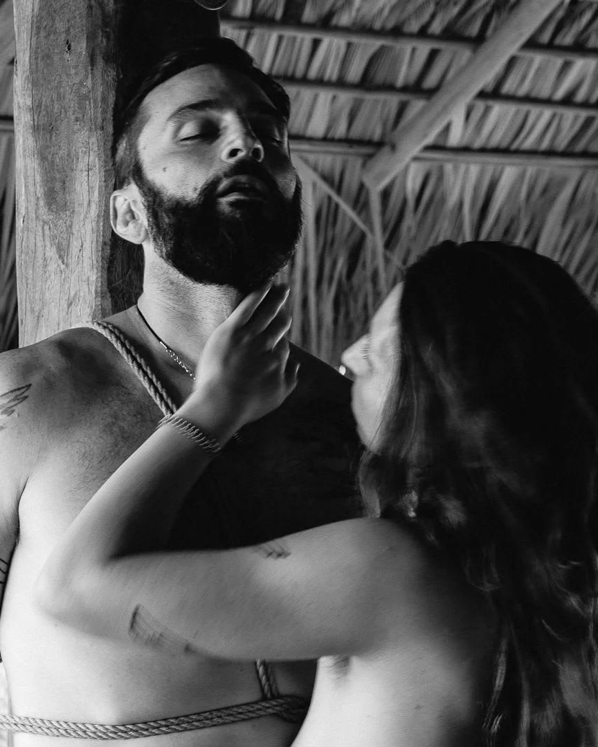 Una pareja en un entorno rústico, la mujer toca la cara del hombre con las manos, ambos en actitud íntima, en un espacio con techo de paja.