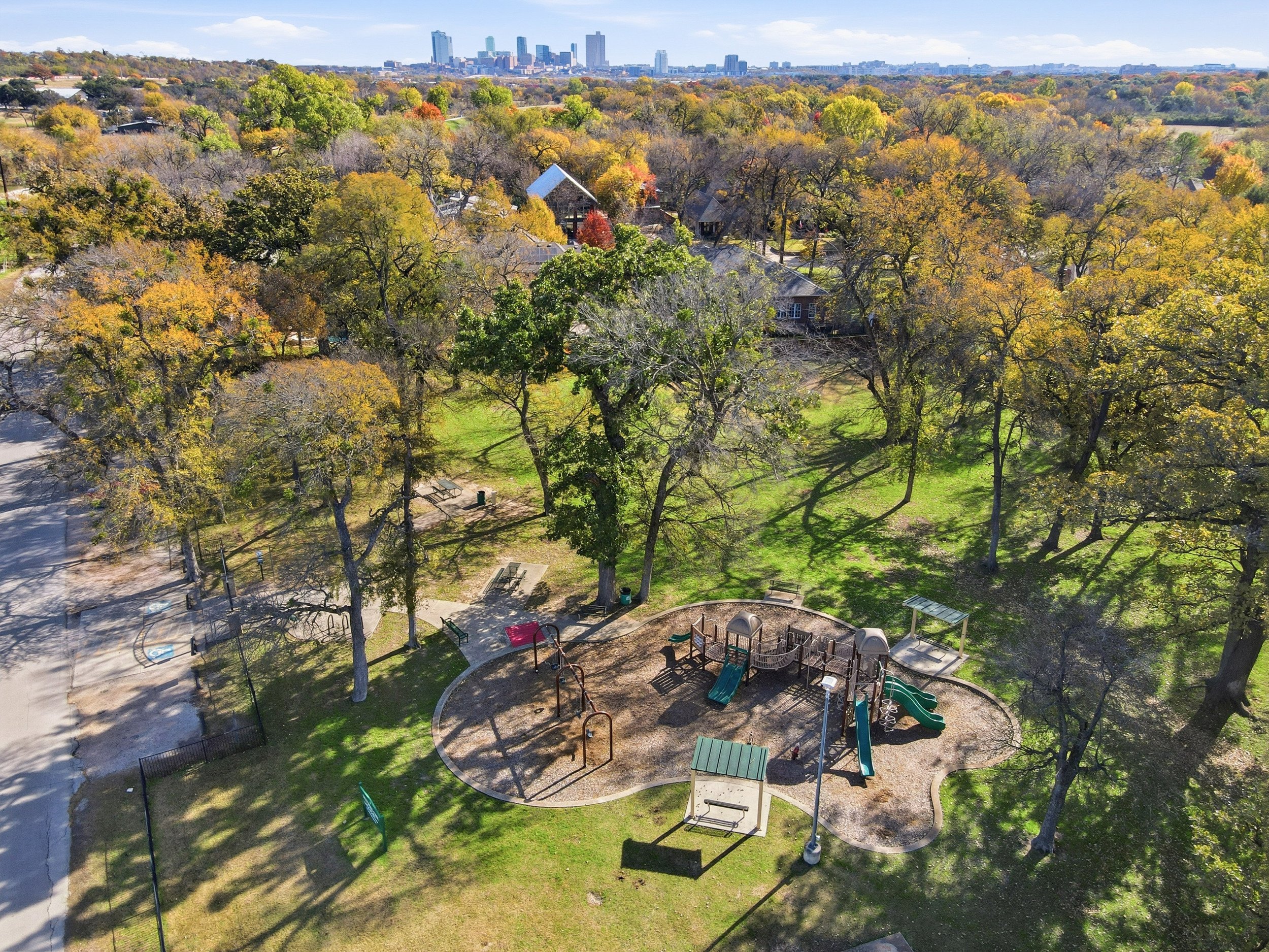 Aerial view of a park with trees and a playground, with a city skyline in the background.