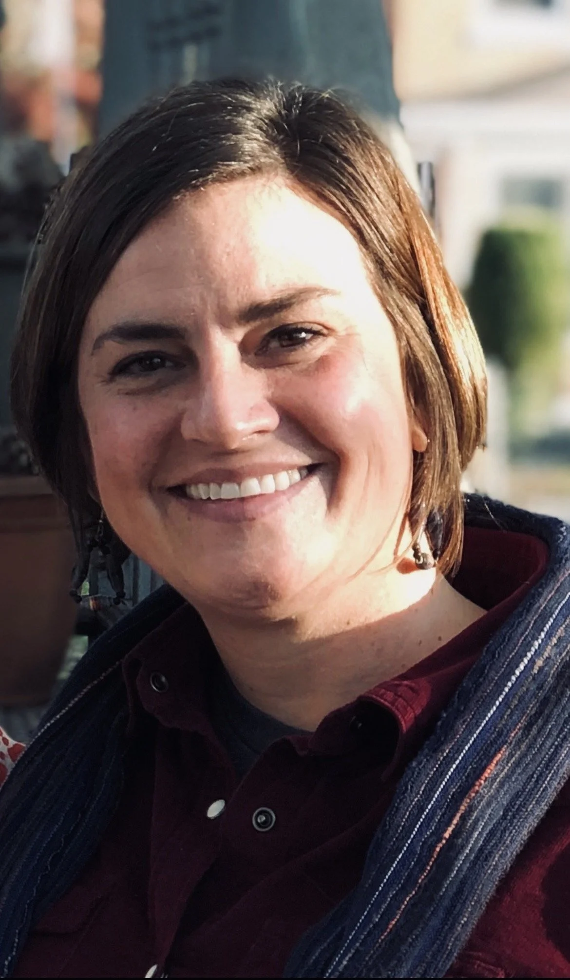 A woman with short brown hair, smiling, wearing a dark maroon shirt and a striped blazer, with earrings, in an indoor setting with blurred background.