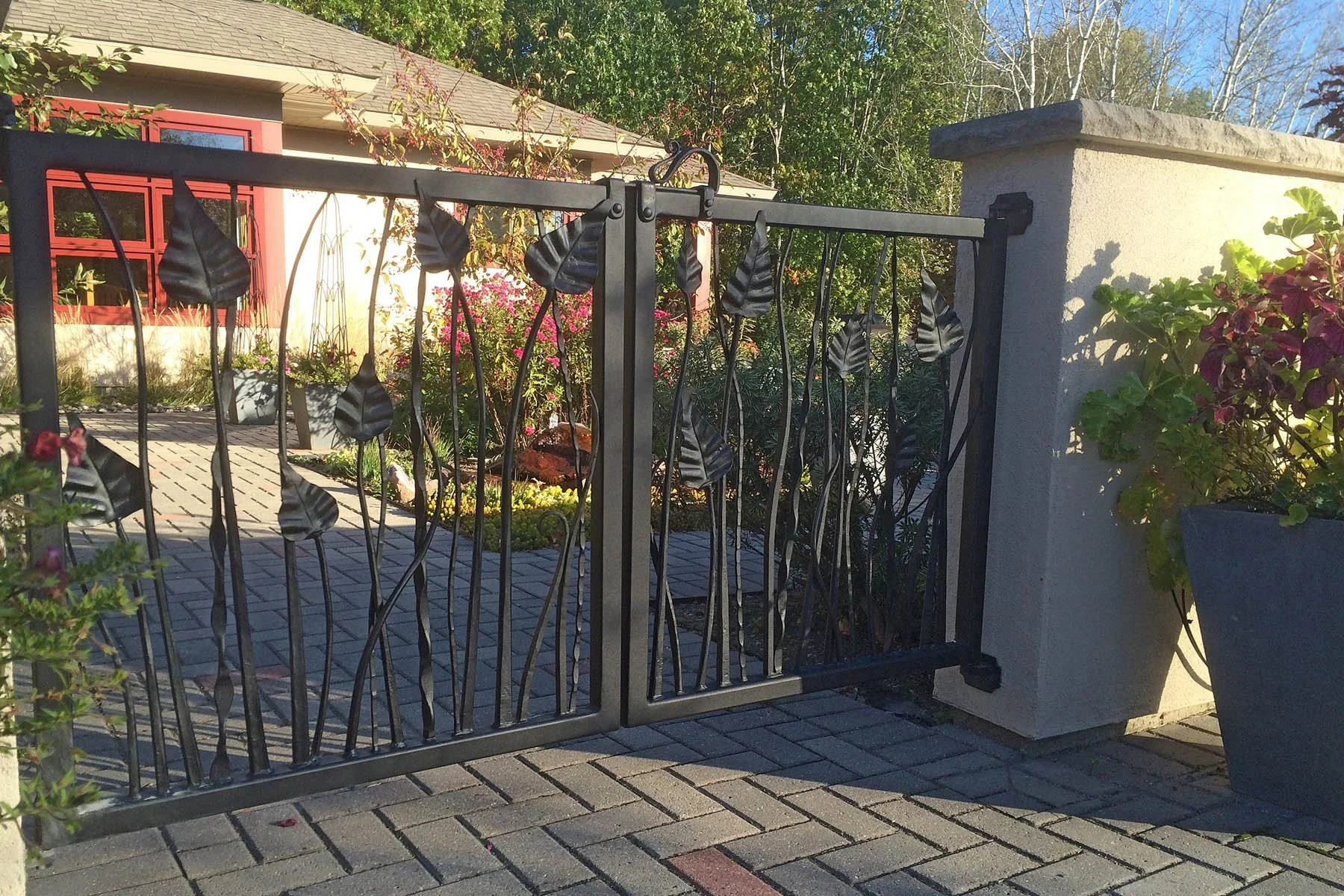 This custom wrought iron garden gate features hand-forged leaf and stem motifs rising organically along slender vertical bars, creating a nature-inspired entrance that blends seamlessly into a lush landscape. Perfect for garden entrances and courtyar