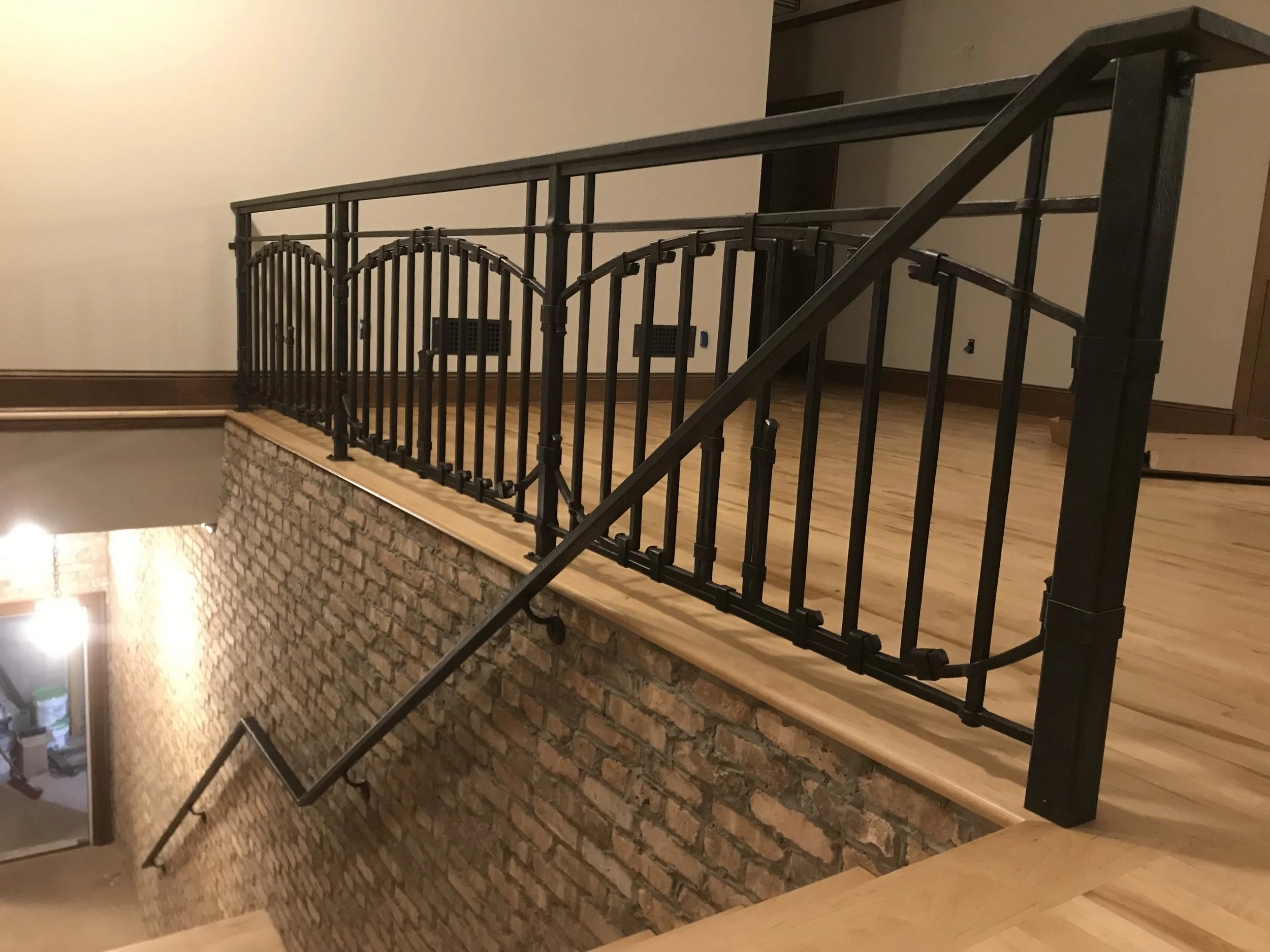 Custom hand-forged wrought iron railing featuring arched paneling and a hammered texture, built by Red Iron Studio for a loft-style stairway landing. This blacksmith-crafted railing pairs beautifully with exposed brick and wide-plank wood flooring, b