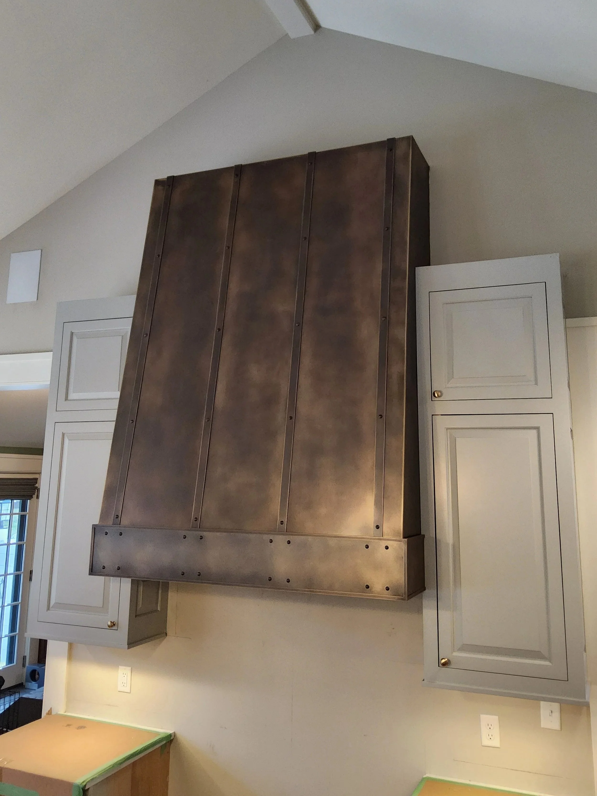 Custom Bronzed Steel Range Hood with Vertical Strapping -This tall, statement range hood features a hand-applied bronzed patina finish with vertical banding and riveted seams, creating a rich, warm focal point beneath a vaulted ceiling. Custom-fabric
