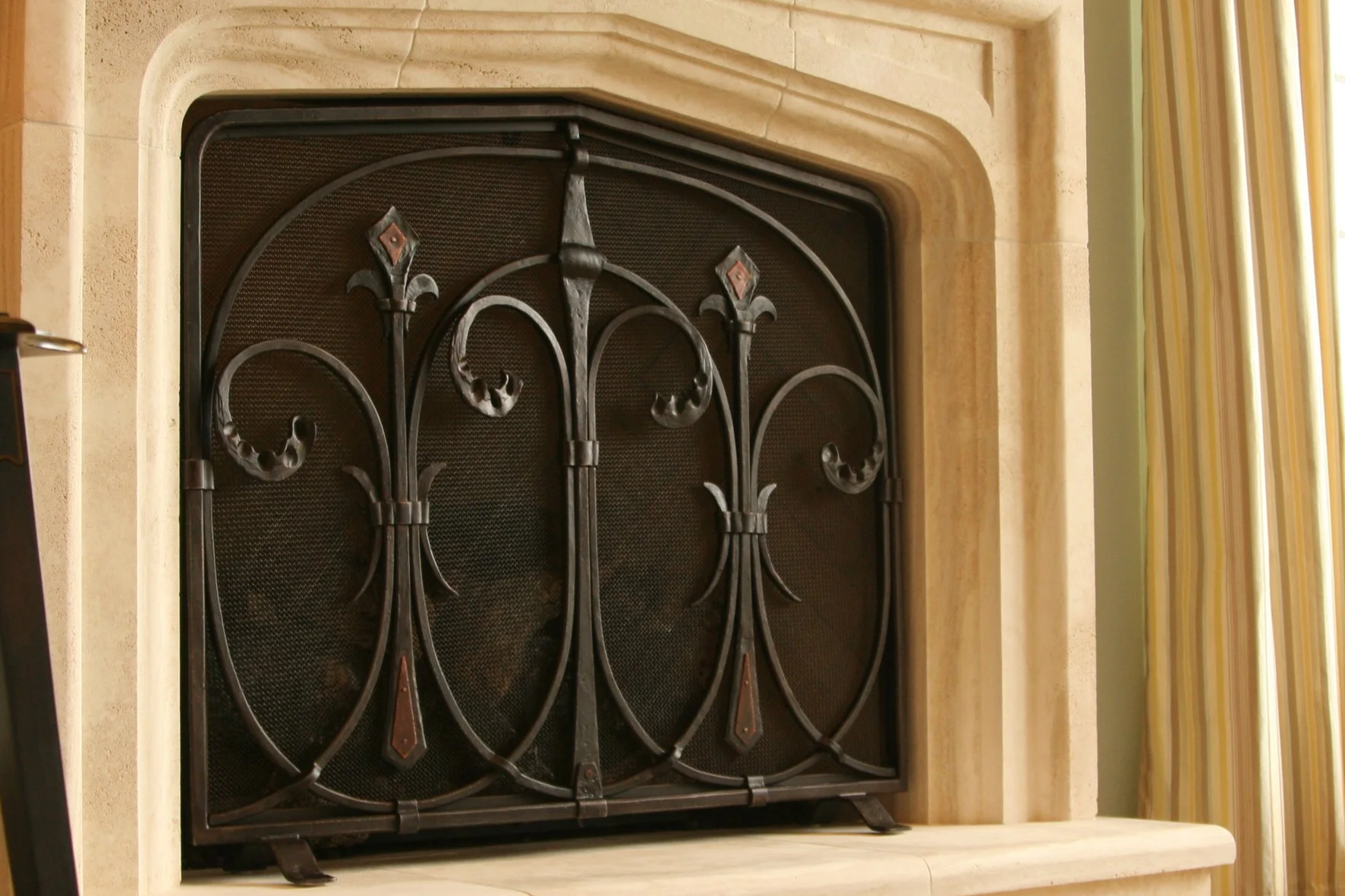 Custom Gothic-inspired wrought iron fireplace screen by Red Iron Studio, featuring hand-forged scrollwork, fleur-de-lis finials, and hammered copper diamond accents, shaped to fit an arched limestone hearth. This decorative fire screen combines tradi
