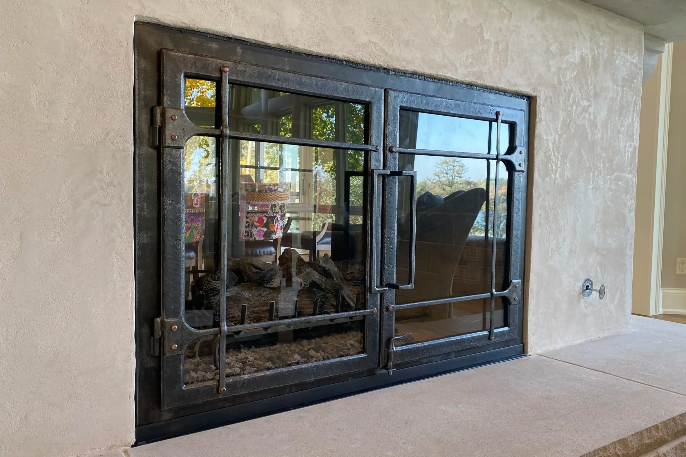 Custom hand-forged fireplace glass doors by Red Iron Studio, showcasing a clean-lined design with hammered iron frames, copper rivet details, and a sturdy forged handle. These handcrafted iron fireplace doors bring a modern-rustic aesthetic to a plas