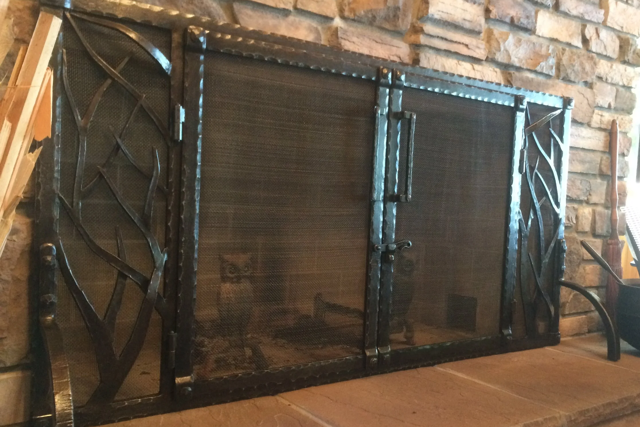 Hand-forged fireplace screen by Red Iron Studio, featuring organic branch-and-twig side panels, a hammered iron frame, and a hand-forged handle set into a natural stone hearth. This rustic fireplace screen showcases nature-inspired blacksmithing, bri