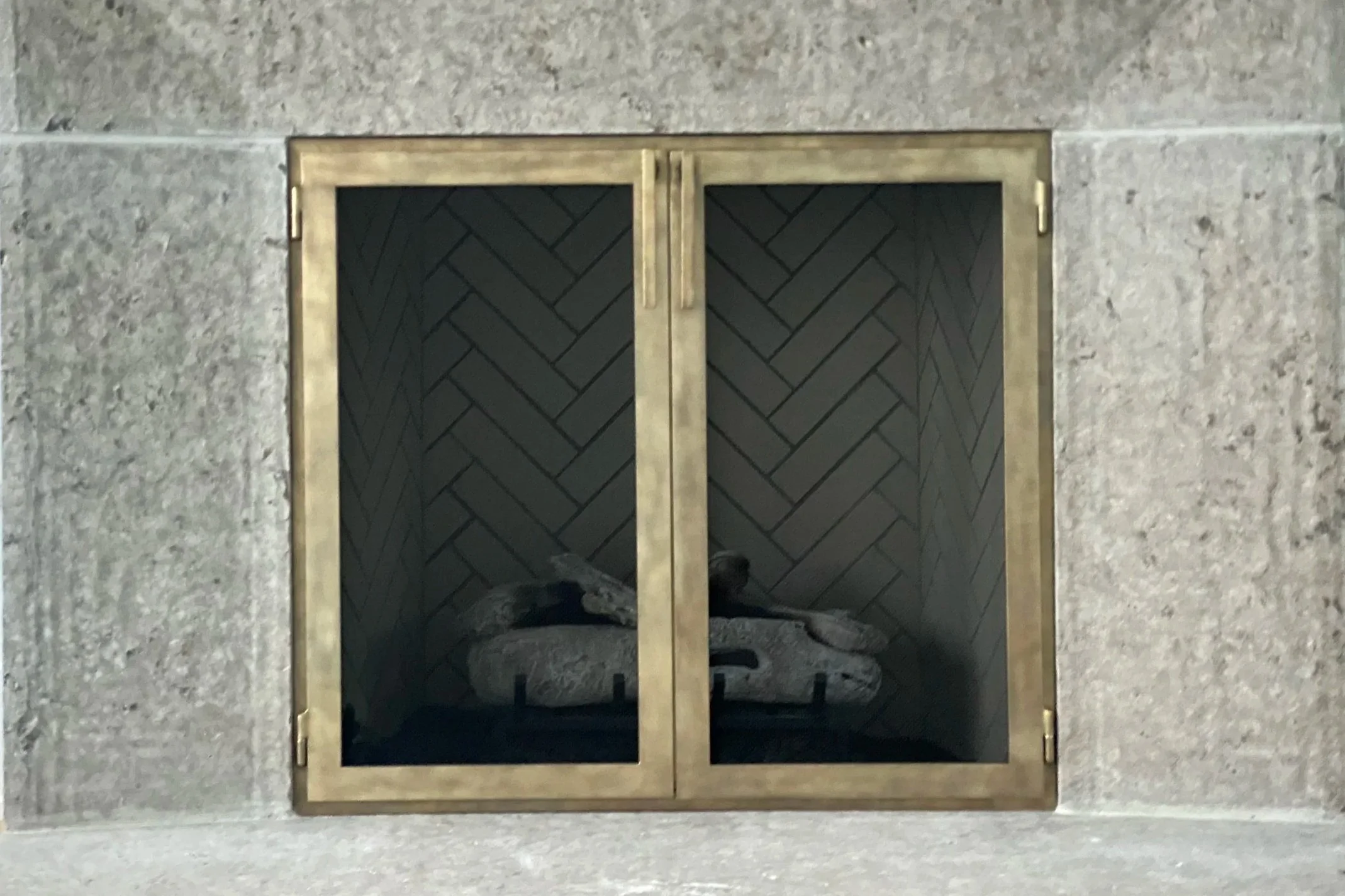 Custom brass fireplace doors by Red Iron Studio, hand-forged with a sleek square frame in a warm antique brass finish to complement a herringbone-tiled firebox and honed stone surround. These handcrafted fireplace doors bring an elegant, modern-luxe 
