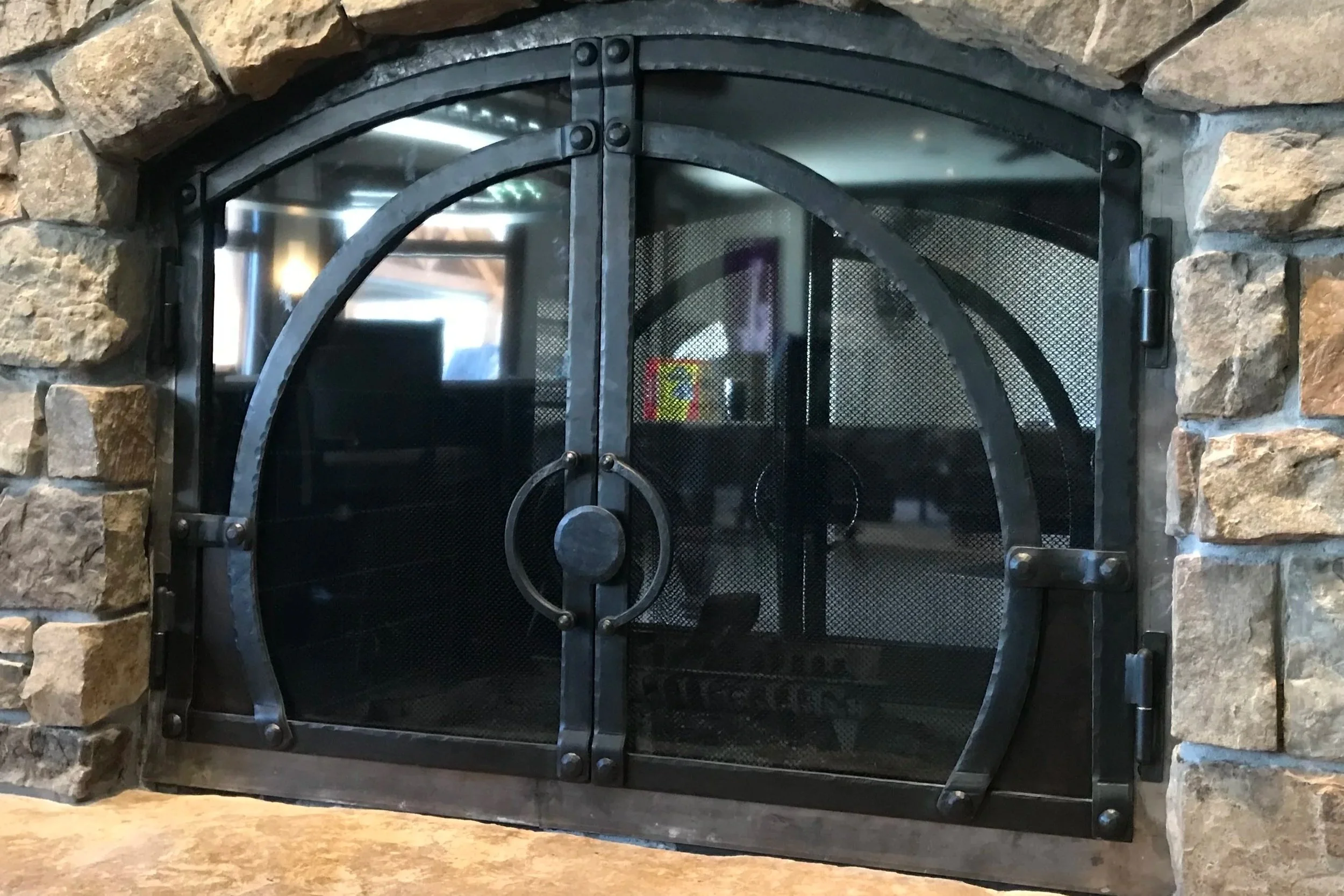 Custom arched fireplace doors by Red Iron Studio, hand-forged in blackened steel with a sweeping curved overlay design and a hand-hammered ring pull handle. These handcrafted iron fireplace doors are built to fit a rugged stone arch, pairing rustic m