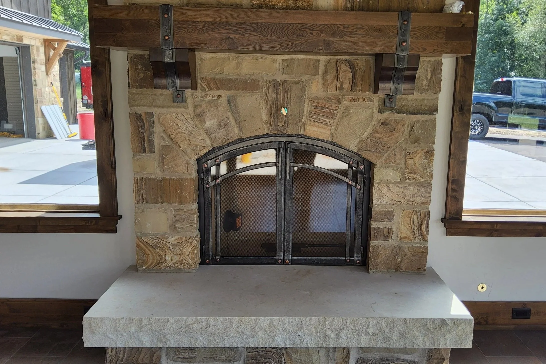 Custom arched fireplace doors by Red Iron Studio, hand-forged with copper rivet details to complement a rustic stone fireplace, hand-hewn wood mantel, and matching corbels. These handcrafted iron fireplace doors add warm, artisan detail to a farmhous