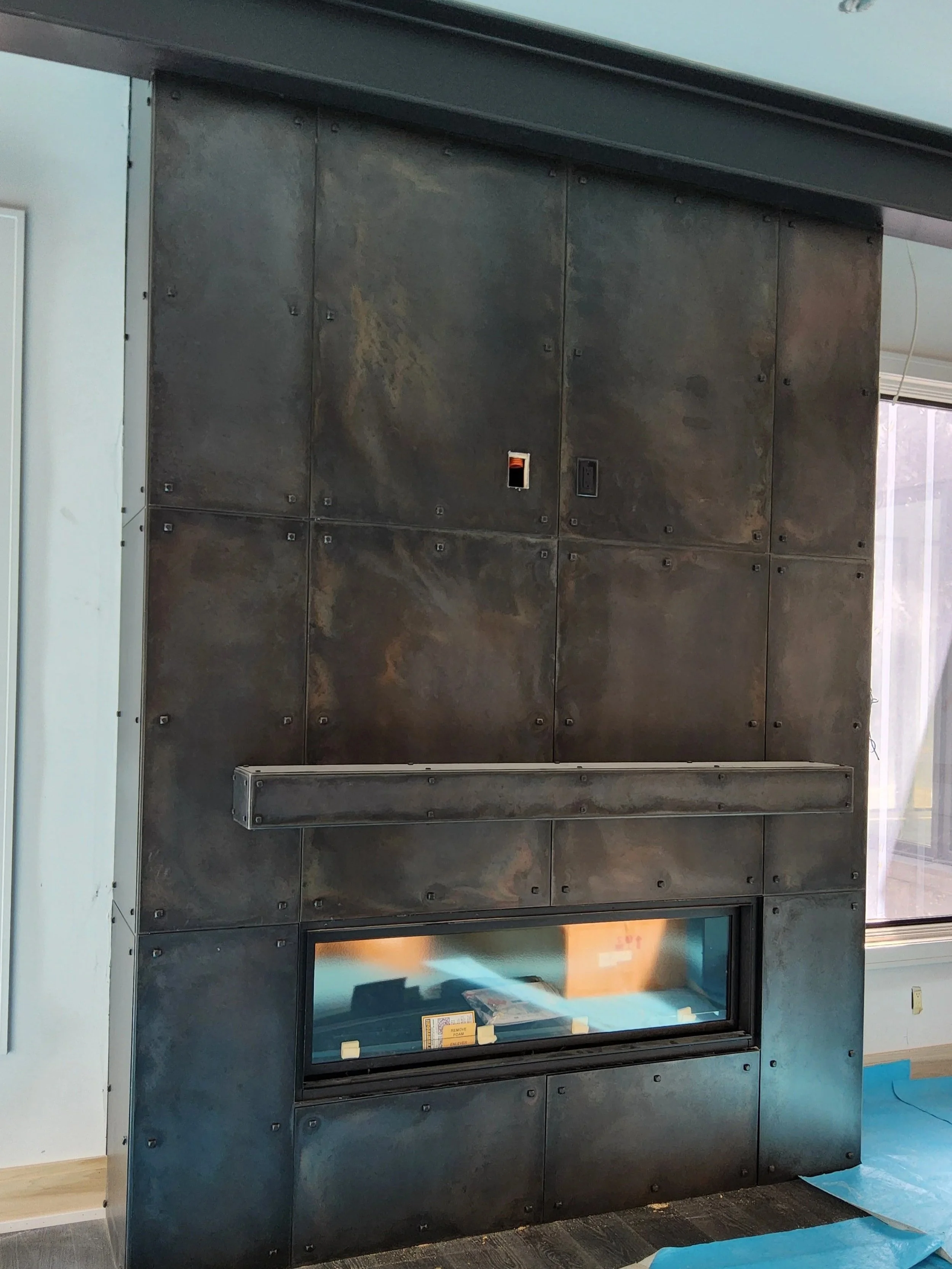 Custom riveted steel fireplace surround by Red Iron Studio, hand-fabricated in a rich blackened patina with a paneled plate design and integrated floating mantel shelf around a linear gas fireplace. This full-height custom fireplace surround delivers