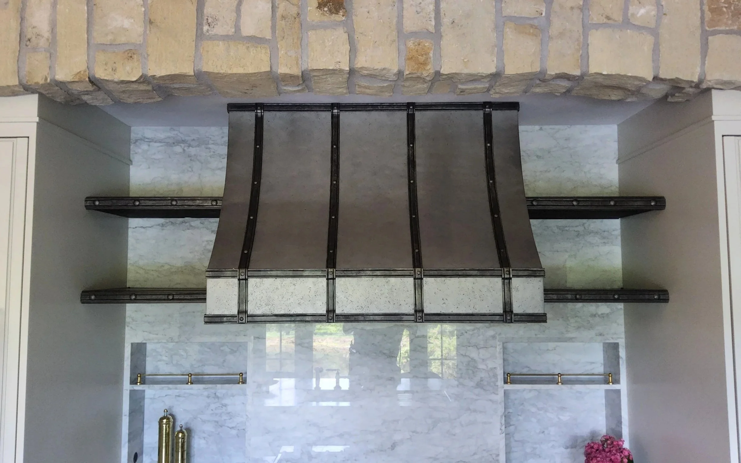 Custom blackened steel range hood with riveted banding and extended iron support straps, set against a marble slab backsplash beneath a stacked stone chimney — handcrafted by Red Iron Studios.