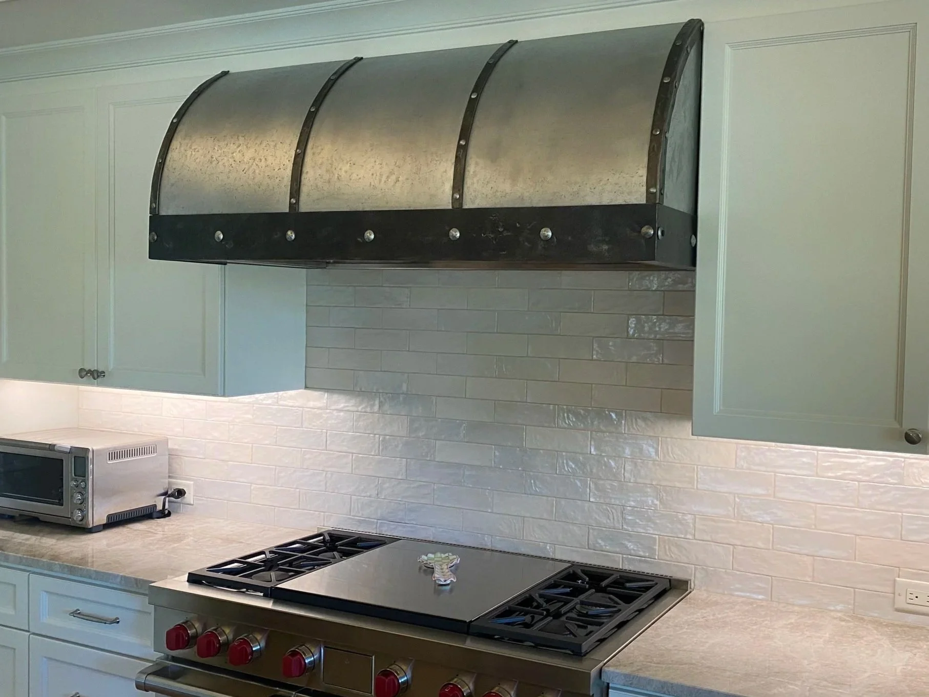 Custom barrel-curved galvanized steel range hood with riveted iron banding above a stainless gas range, set against a white subway tile backsplash and sage green cabinetry — handcrafted by Red Iron Studios.