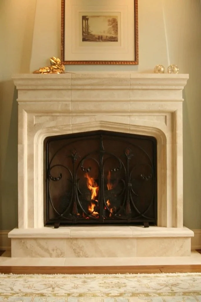 Custom Gothic-inspired wrought iron fireplace screen by Red Iron Studio, featuring hand-forged scrollwork, fleur-de-lis finials, and hammered copper diamond accents, shown glowing beautifully in front of an arched limestone hearth. This decorative fi