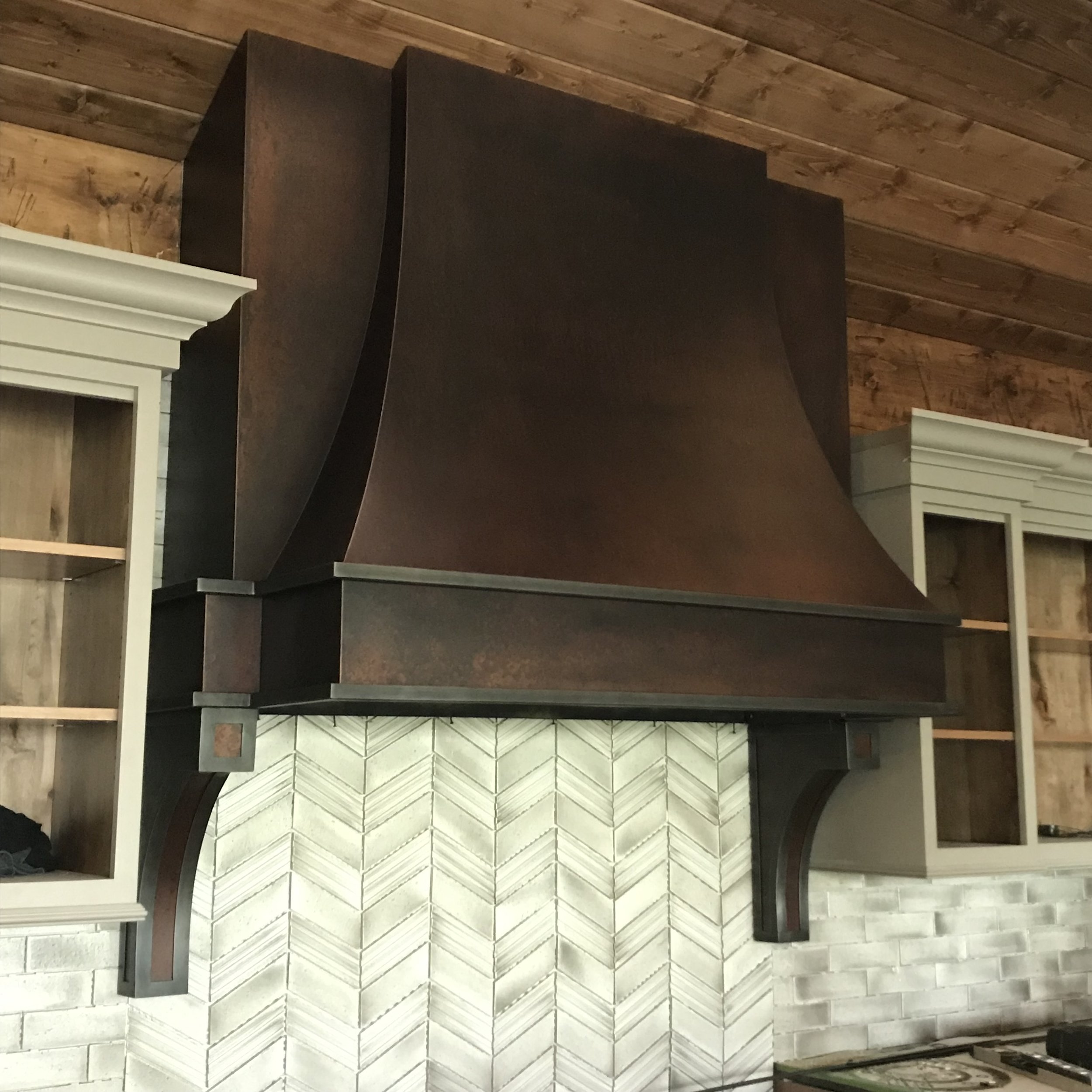 Custom Aged Copper Range Hood with Decorative Iron Corbels -This dramatic, draped-panel range hood showcases a rich aged copper finish paired with hand-forged iron corbels and trim, creating a bold architectural centerpiece for a rustic luxury kitche