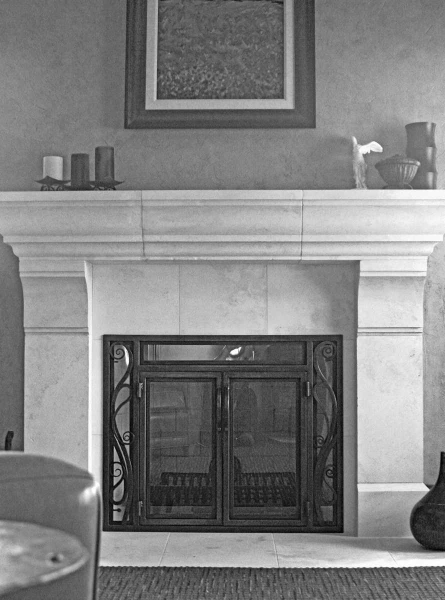 Custom fireplace doors by Red Iron Studio, hand-forged with elegant scrolled iron side panels, divided glass, and a transom-style top window to fit a classic stone mantel. These handcrafted iron fireplace doors combine traditional scrollwork with cle